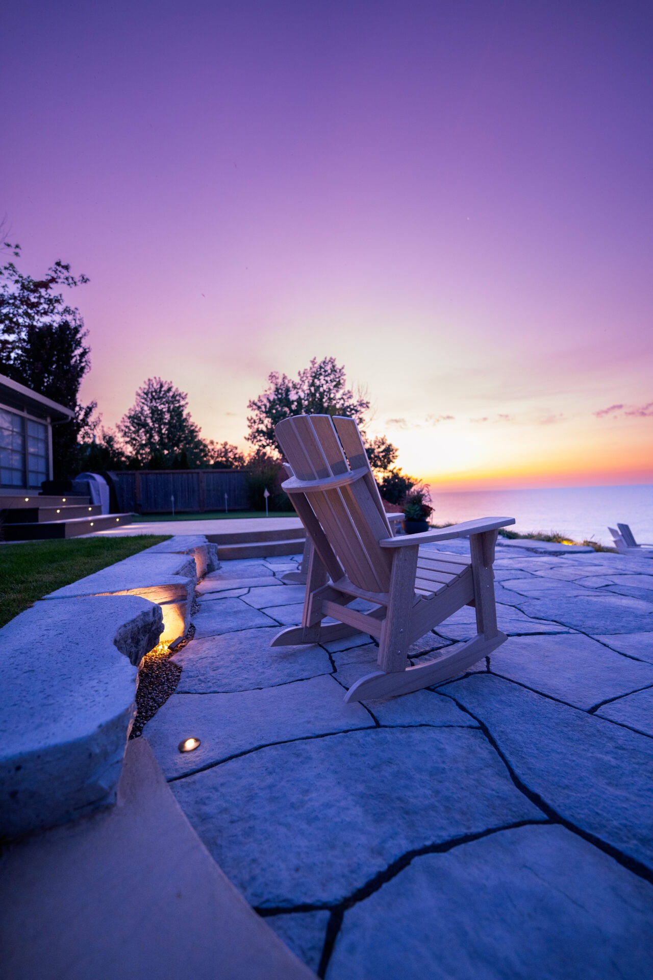 An outdoor scene at dusk with a wooden chair on a stone patio, overlooking a calm sea with a purple and orange gradient sky.