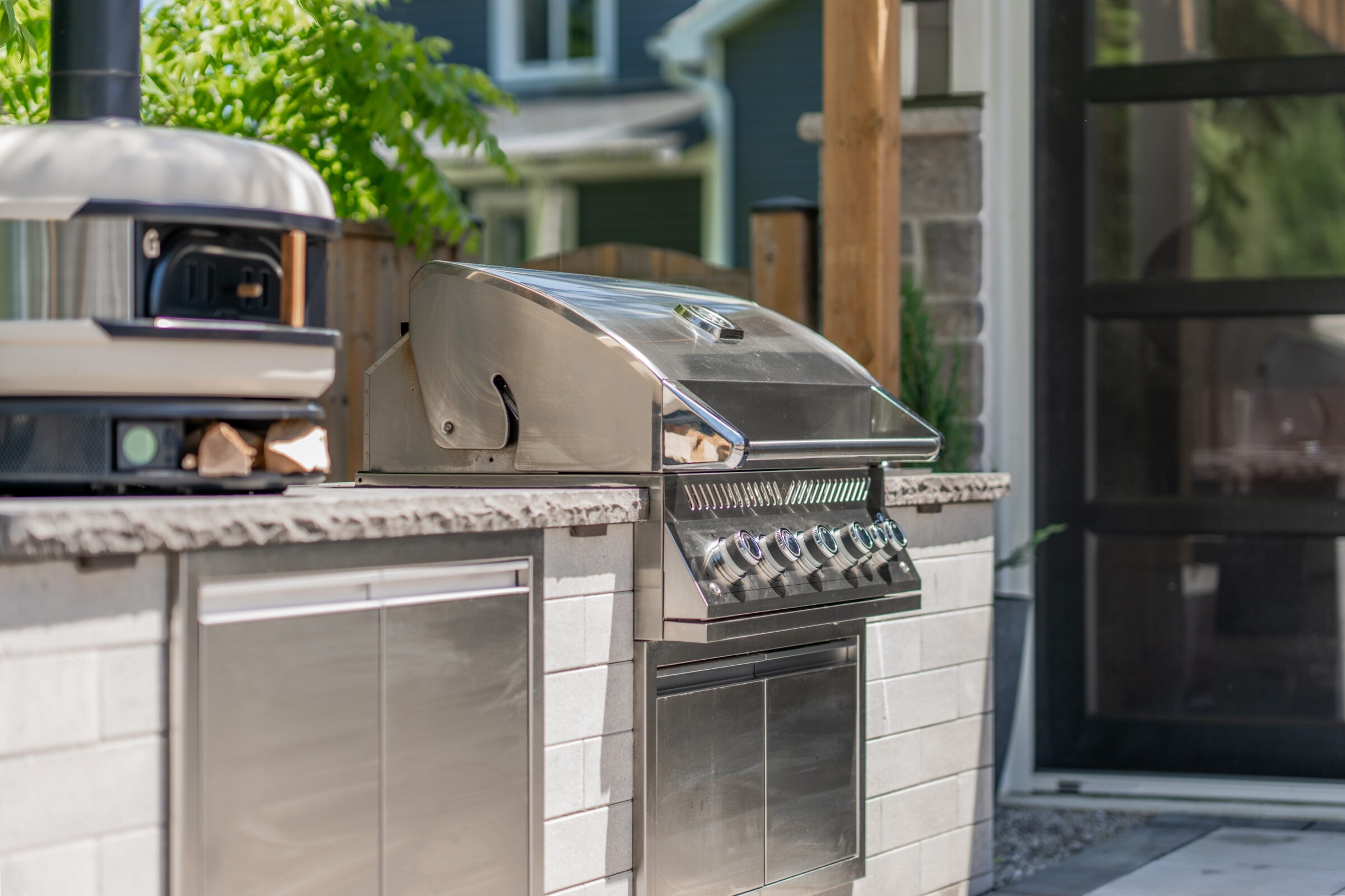 An outdoor kitchen with a stainless steel gas grill, built into a stone countertop, near a pizza oven, with a view of trees and a house.