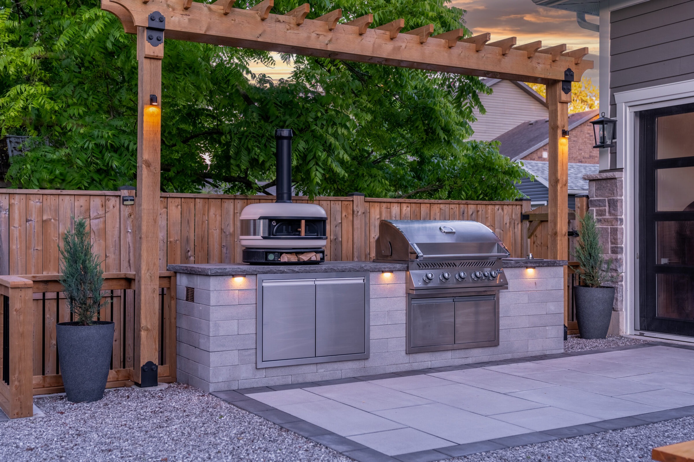 A well-equipped outdoor kitchen on a patio with a built-in grill, pizza oven, ample counter space, and lighting, enclosed by a wooden fence and pergola.