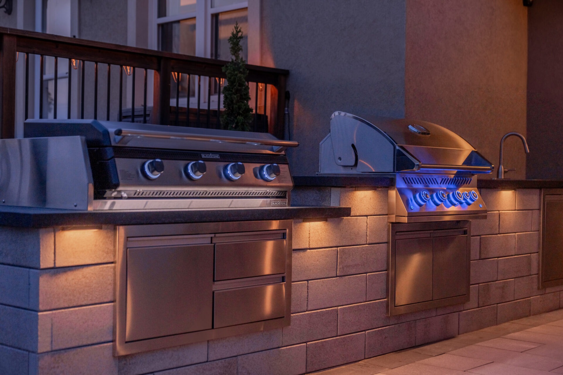 An outdoor kitchen setup with stainless steel grills, under-cabinet lighting, stone countertops, and a wooden railing in a twilight setting.