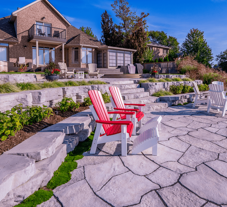 This image shows a serene backyard with red and white Adirondack chairs, stone landscaping, lush plants, and a beautiful house with a balcony.