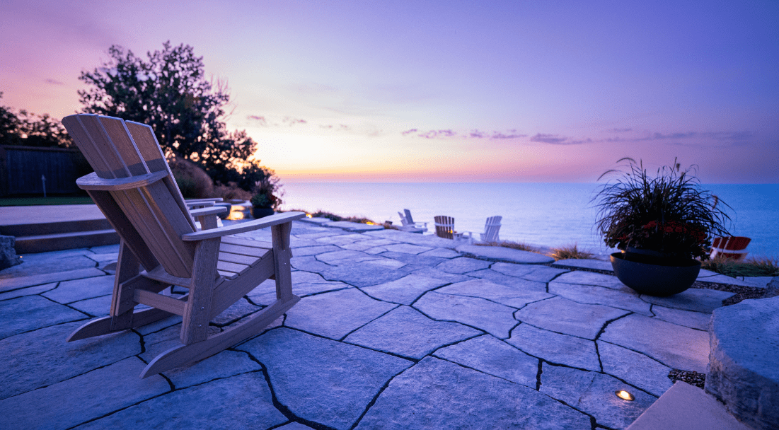 An outdoor patio with stone flooring, Adirondack chairs, and potted plants overlooks a calm sea during sunset, with vibrant colors in the sky.