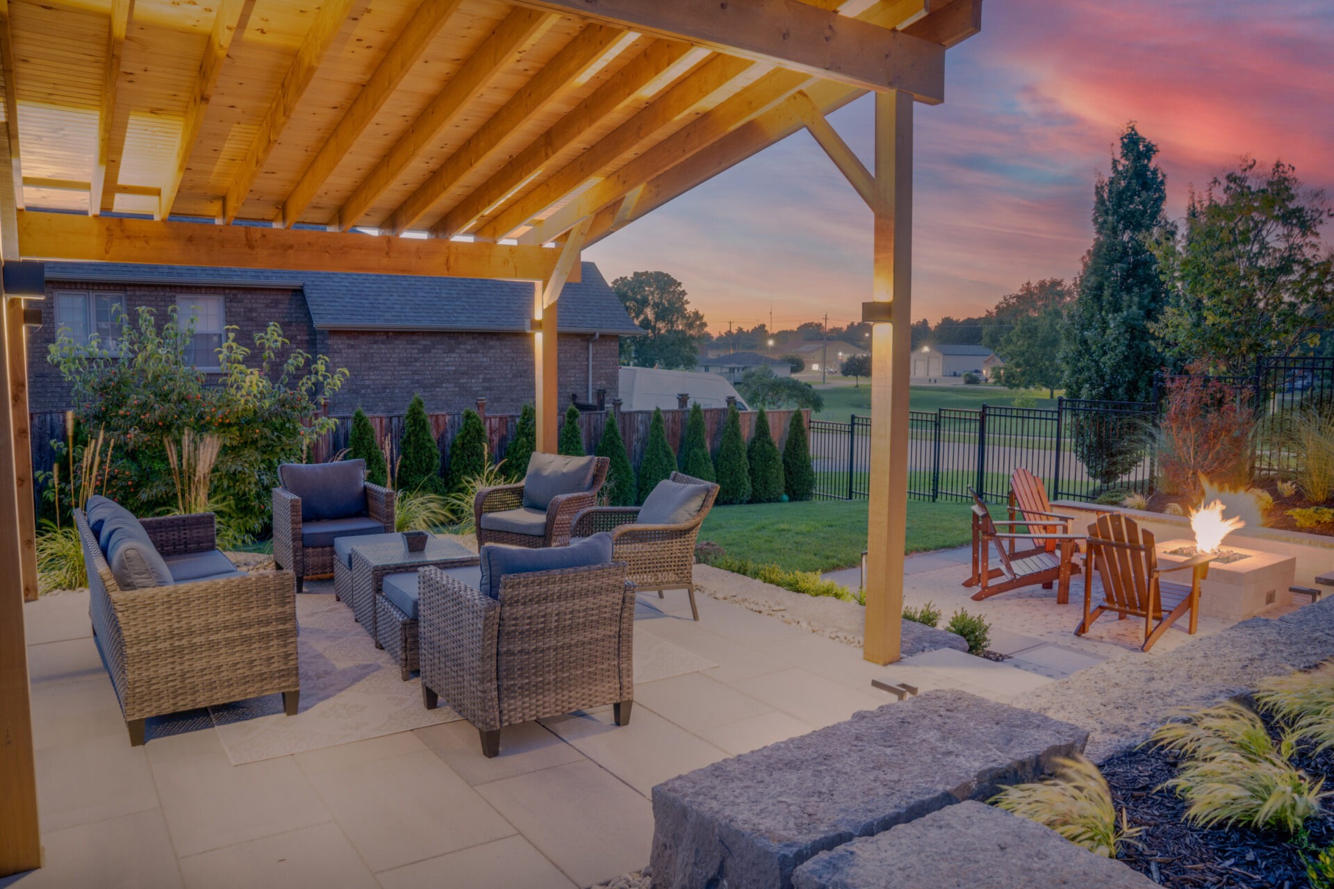 An outdoor patio with wicker furniture under a wooden pergola. A fire pit glows warmly during dusk, with a landscaped and fenced backyard visible.