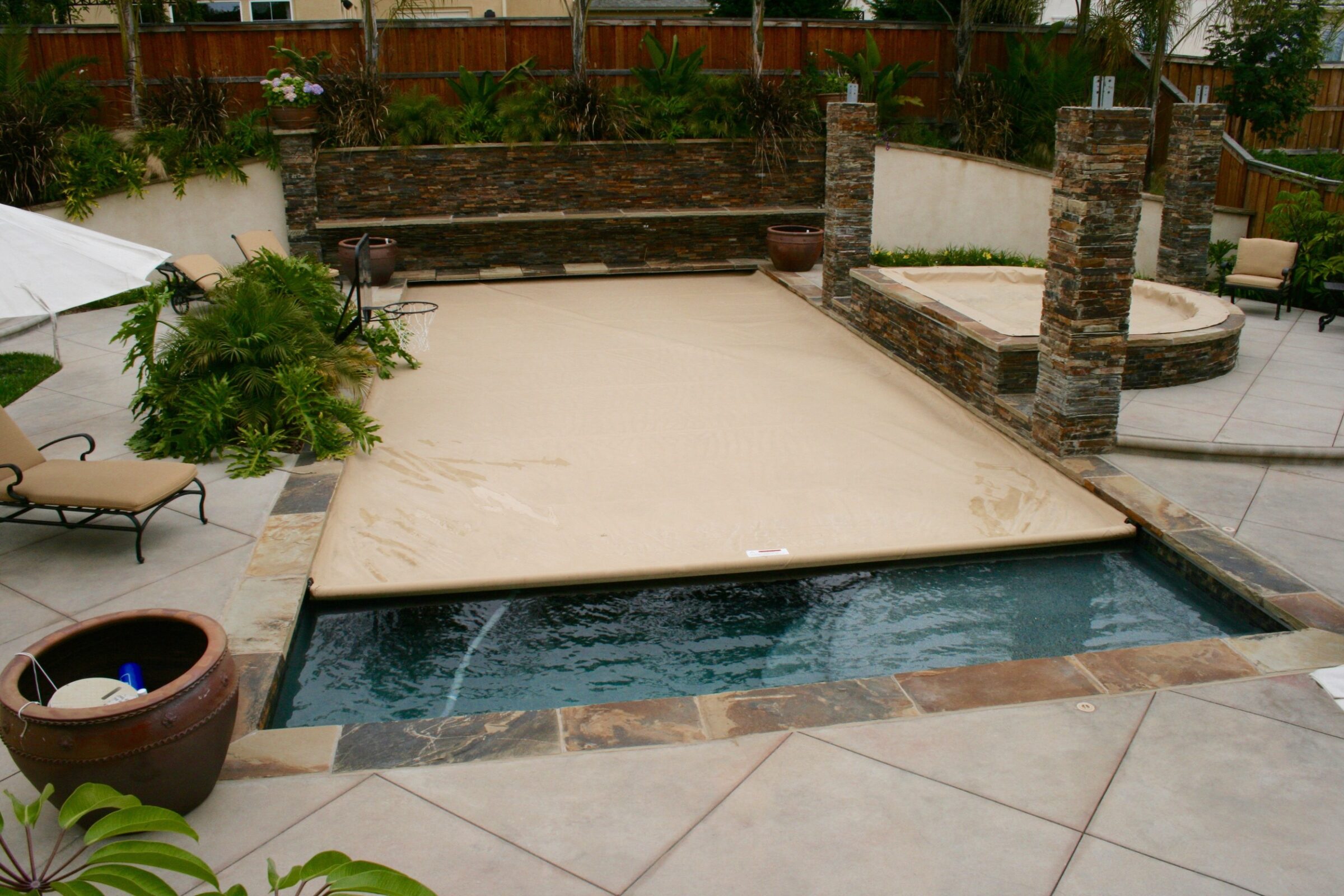 This image shows a backyard with a covered swimming pool, stone benches, potted plants, sun loungers, and a lawn under overcast skies.