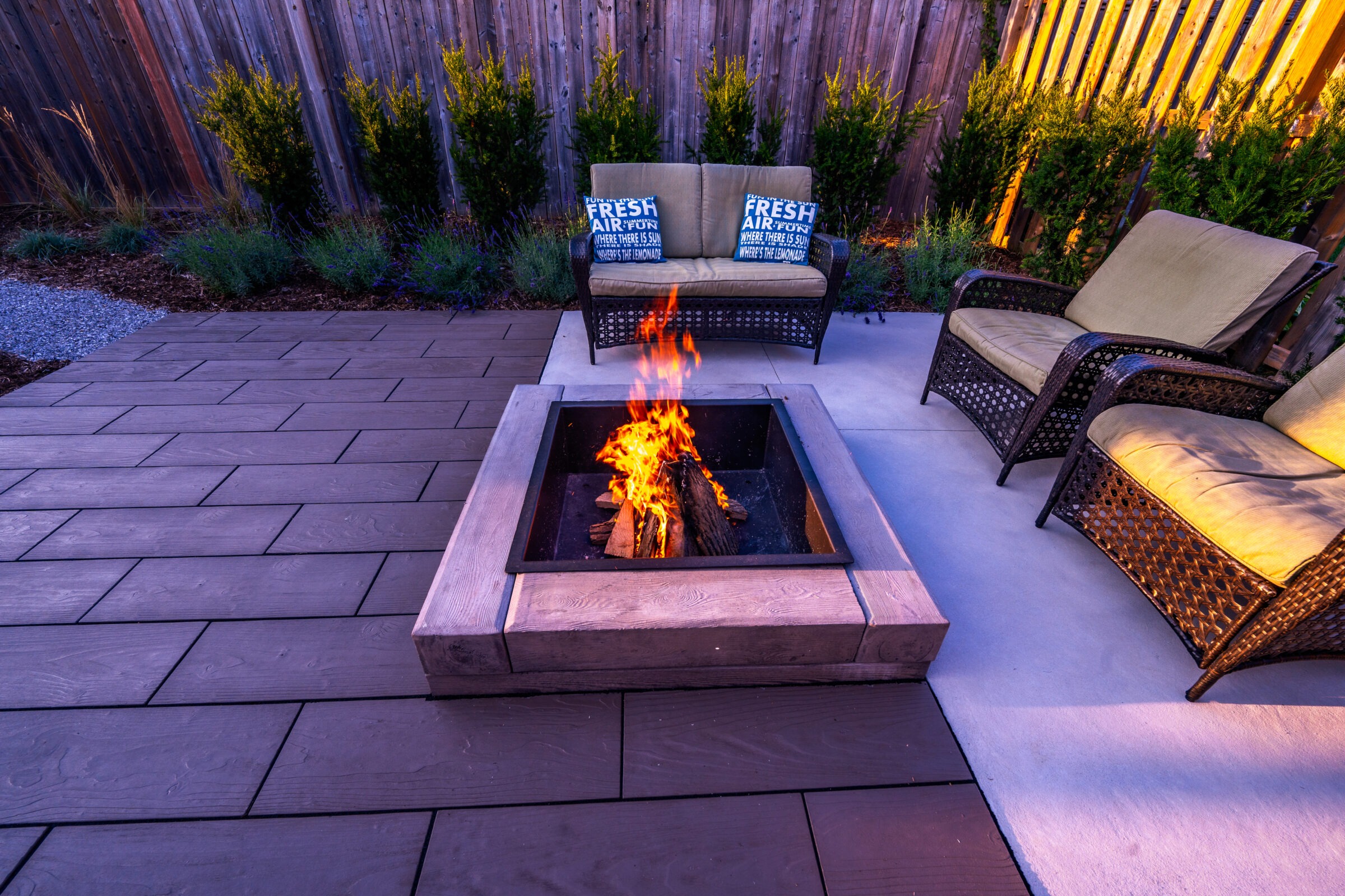 An outdoor patio with a fire pit in the center, surrounded by wicker furniture with cushions, and a wooden fence with shrubs in the background.