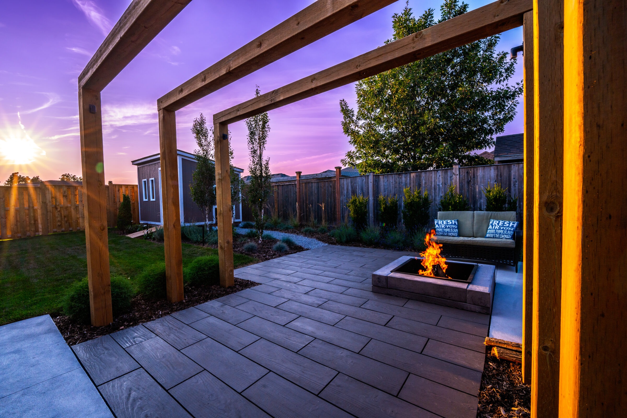 A tranquil backyard at sunset with a wooden pergola, fire pit, bench with pillows, grass, trees, and a wooden fence. The sky is colorful.