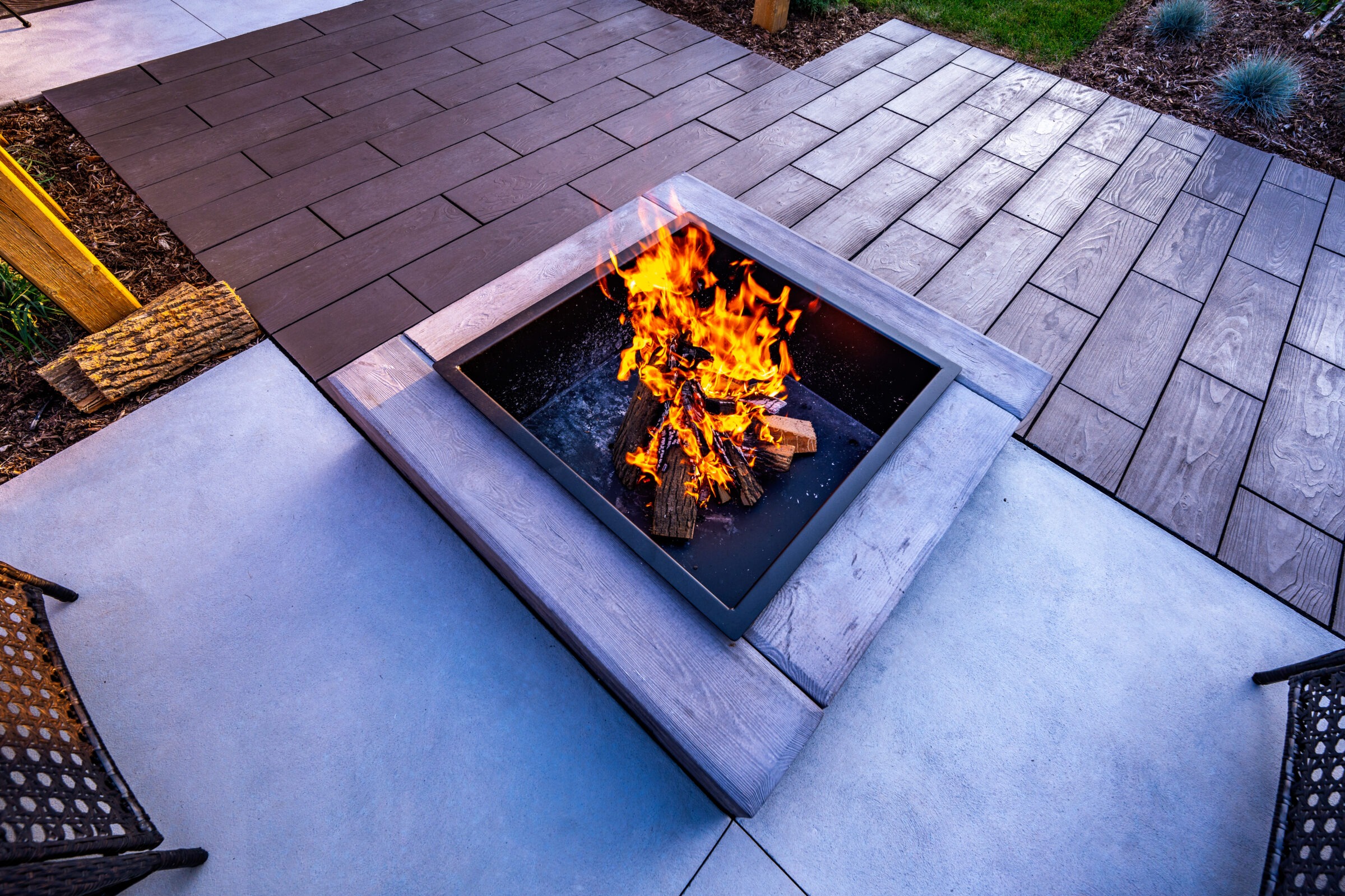 Outdoor setting with a fire pit blazing surrounded by a contrasting pattern of pavers. Comfortable chairs nearby suggest a relaxing entertainment area.