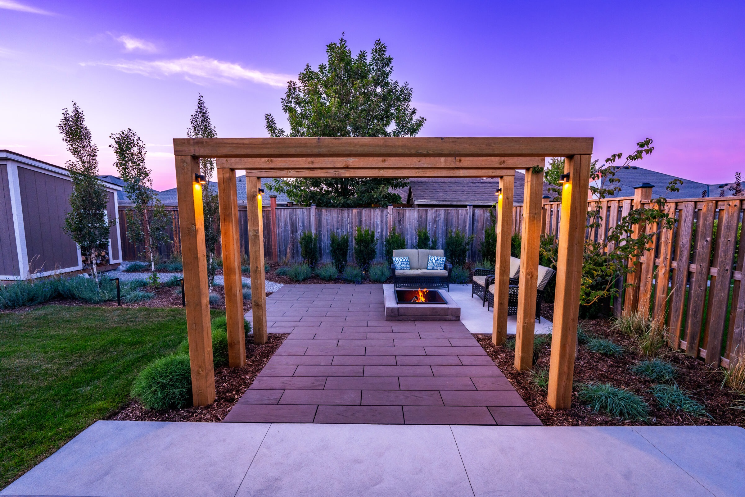 A tranquil outdoor space at dusk with a fire pit, seating, wooden pergola, pathway, ambient lighting, green lawn, and a wooden fence.