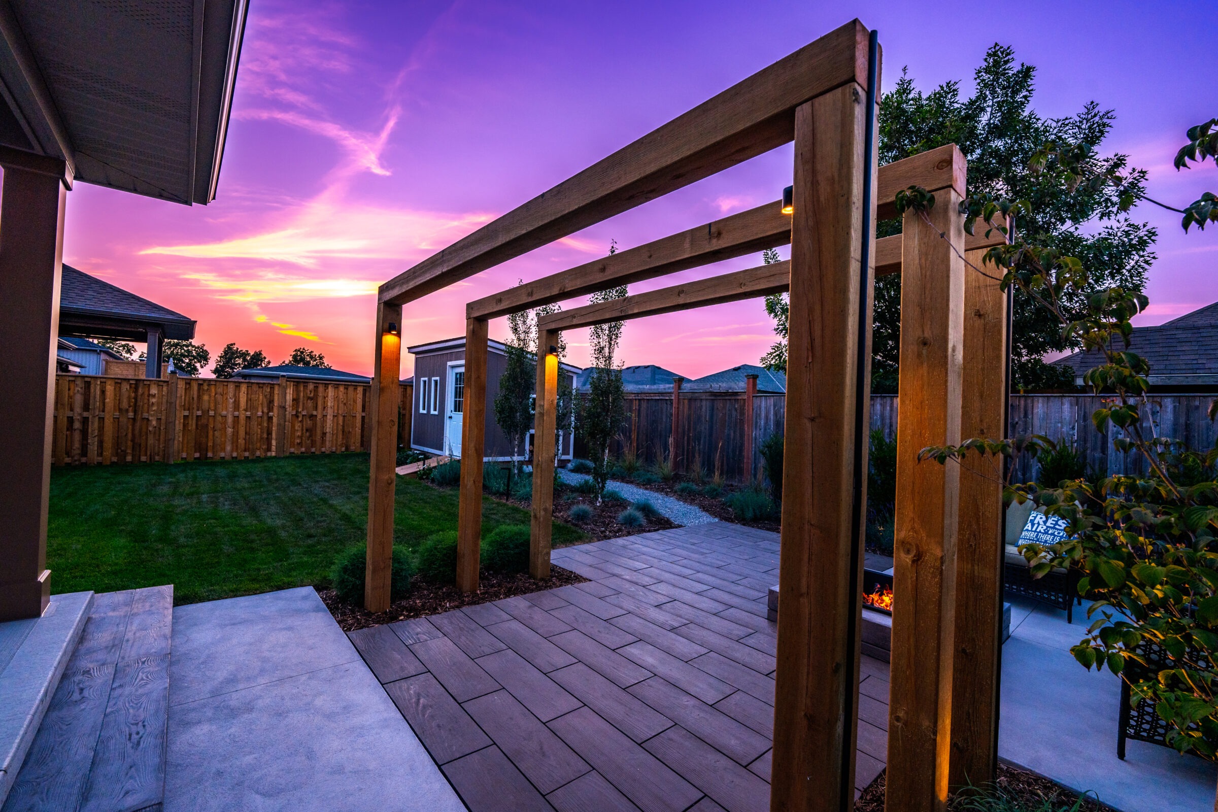 A wooden pergola frames a backyard with a vibrant sunset sky. Lush grass, a fence, and pathway create a tranquil outdoor setting.