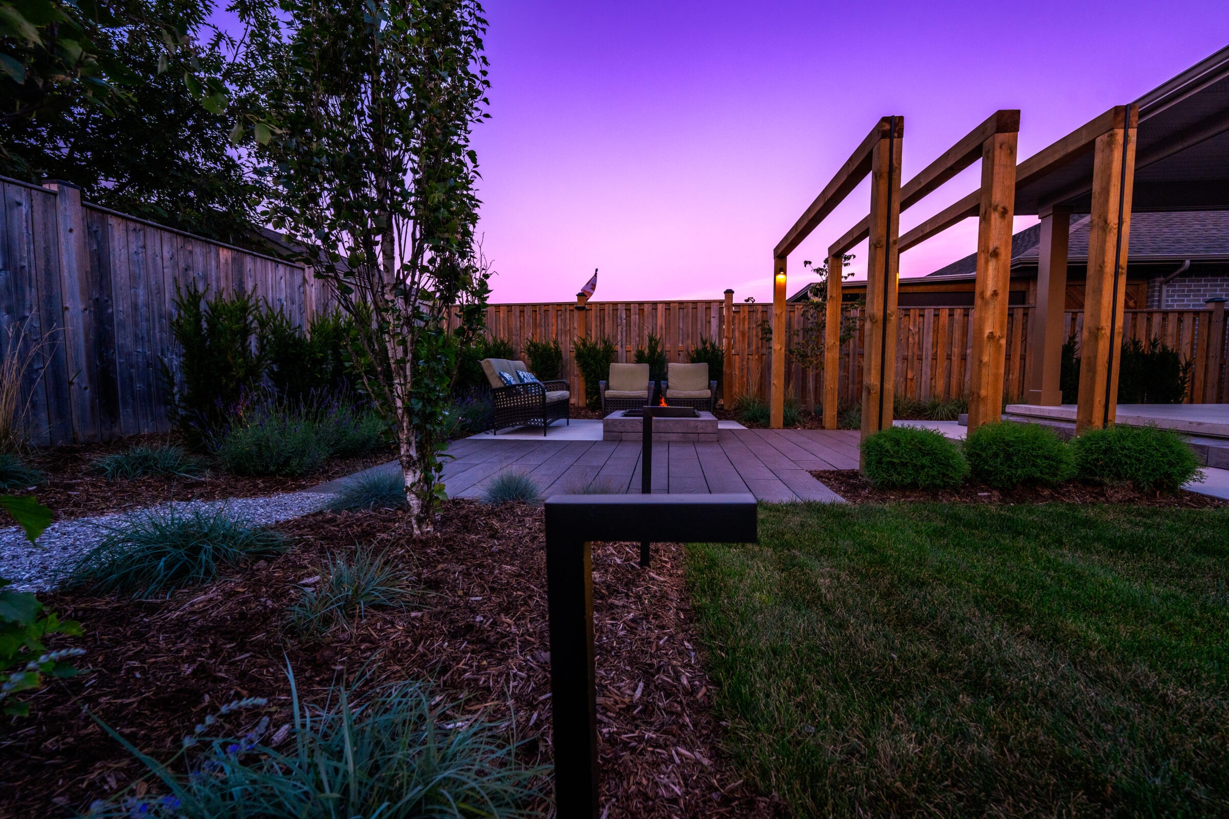 This image depicts a residential backyard at twilight featuring a patio with outdoor furniture, landscaping with greenery, a wooden pergola, and a fence.