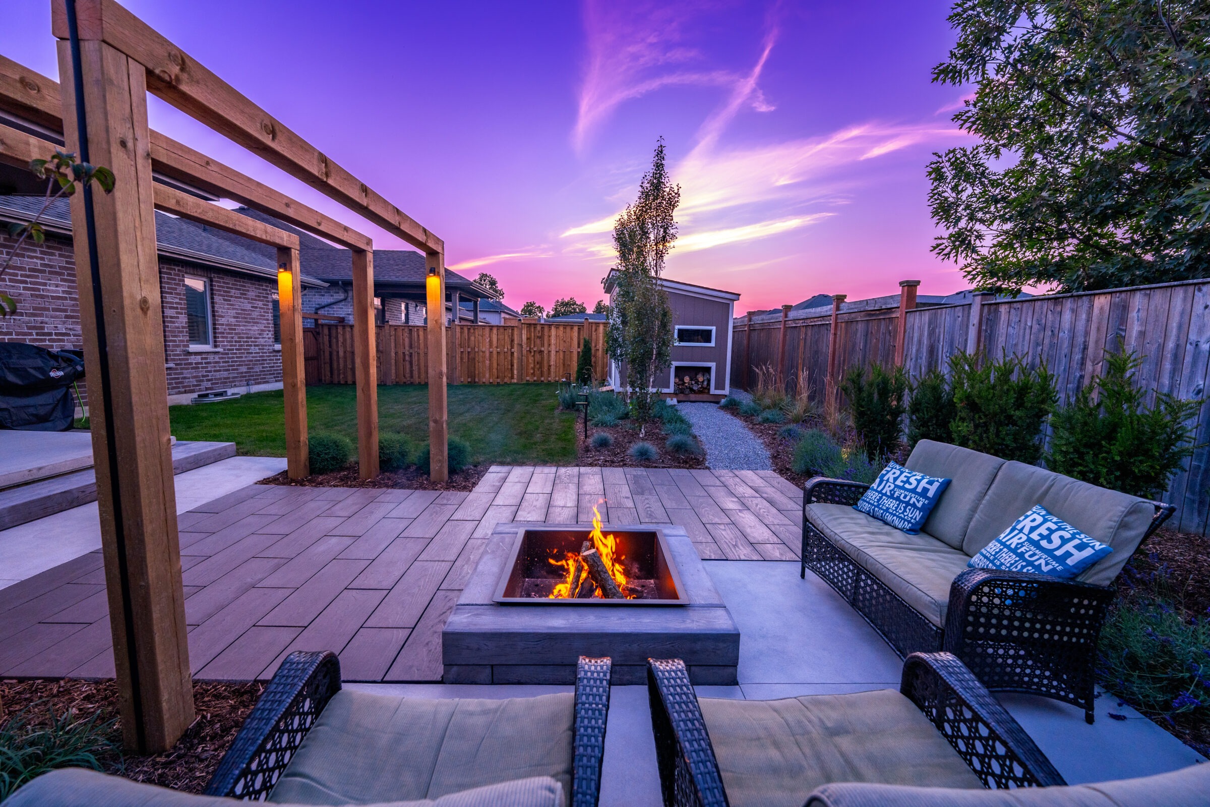 A cozy backyard during sunset with a fire pit, seating area, wooden pergola, and lush landscaping against a vibrant purple sky.