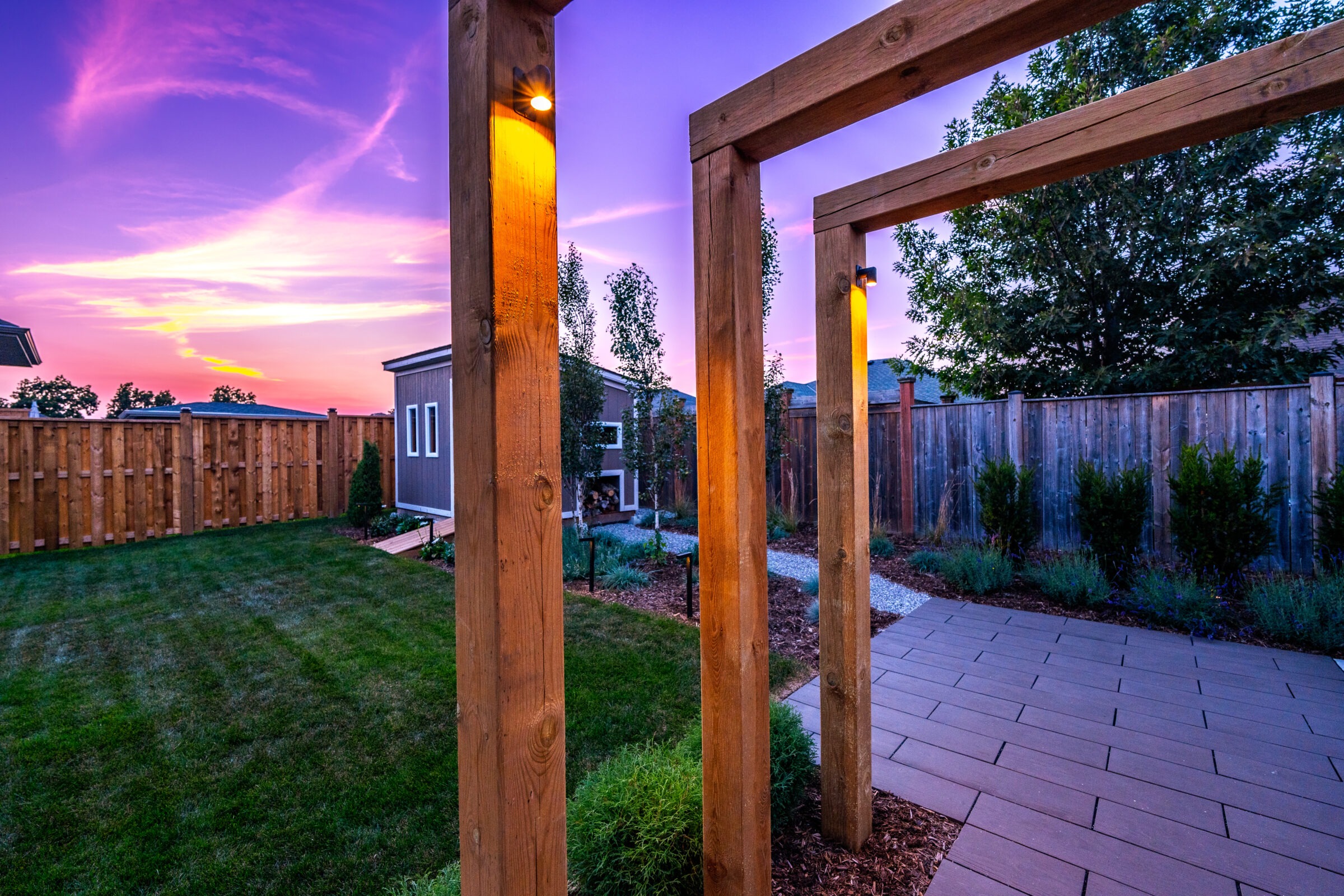 A tranquil backyard with a wooden pergola, manicured grass, a stone pathway, and a wooden fence under a vivid, colorful sunset sky.