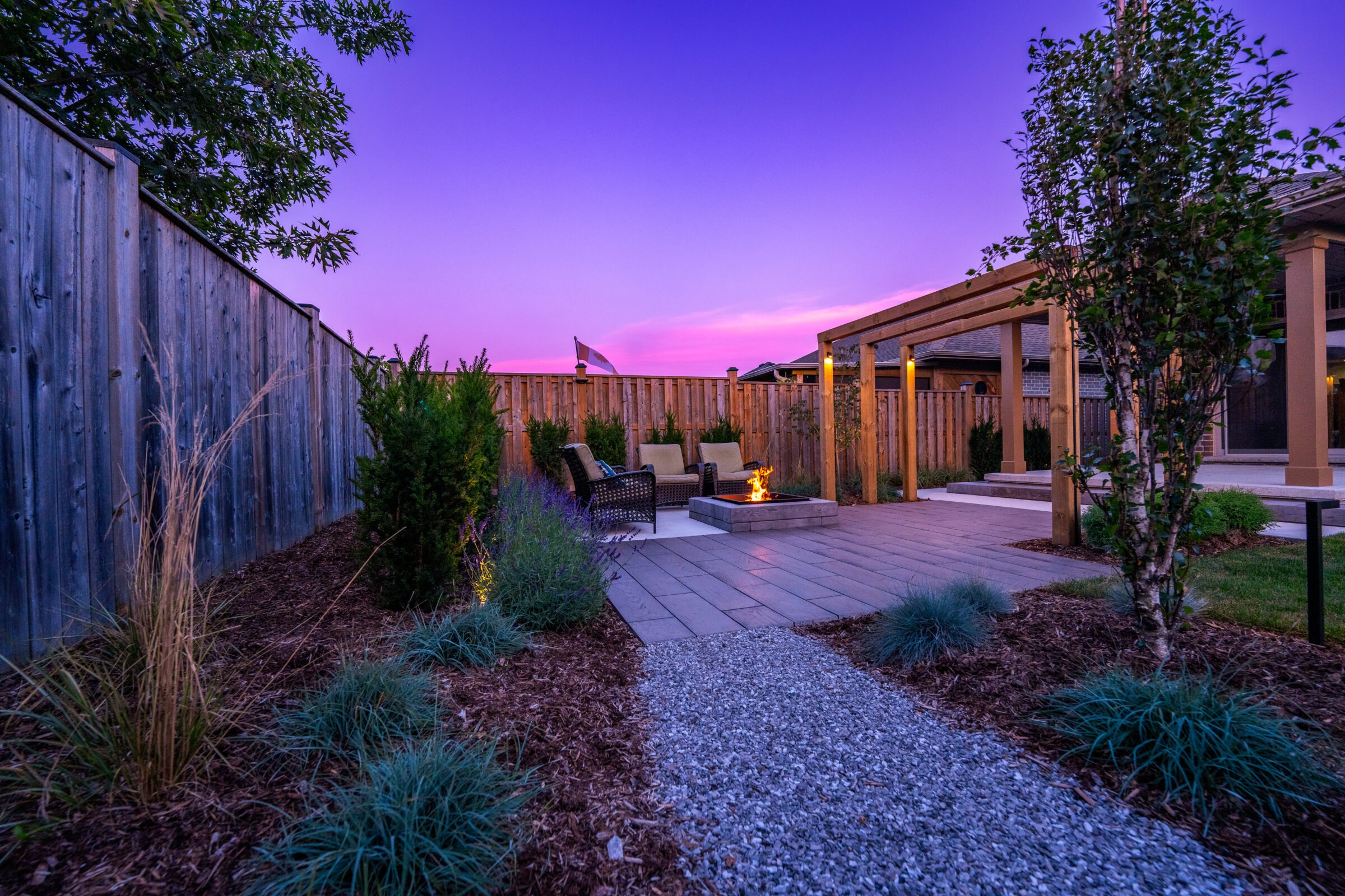 A well-manicured backyard at twilight features a fire pit, seating area, wooden fence, landscaping, and a pergola, under a vibrant purple sky.