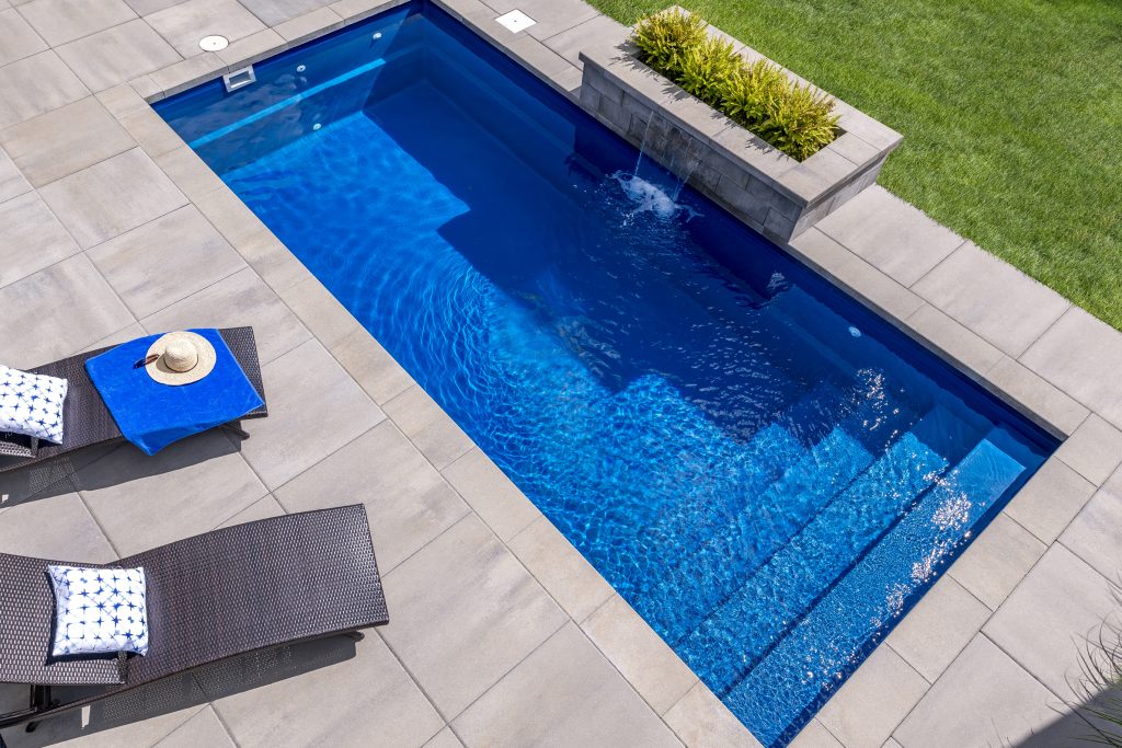 Aerial view of a rectangular backyard swimming pool with sun loungers, pillows, and a straw hat on a tile deck, beside a grassy area.