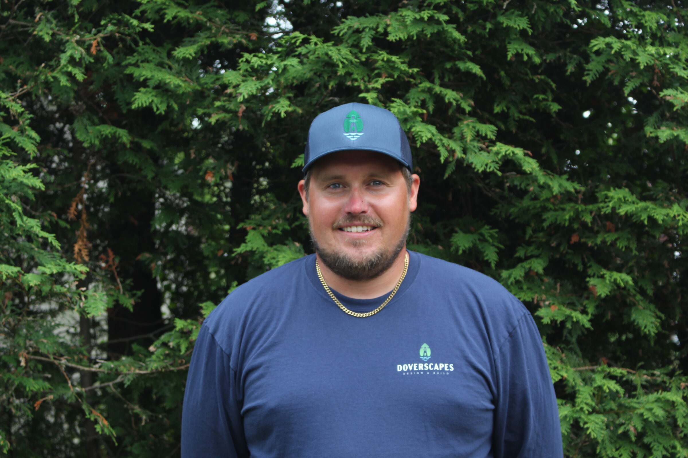A person wearing a dark blue t-shirt and baseball cap smiles, standing in front of dense green foliage. They appear casual and approachable.