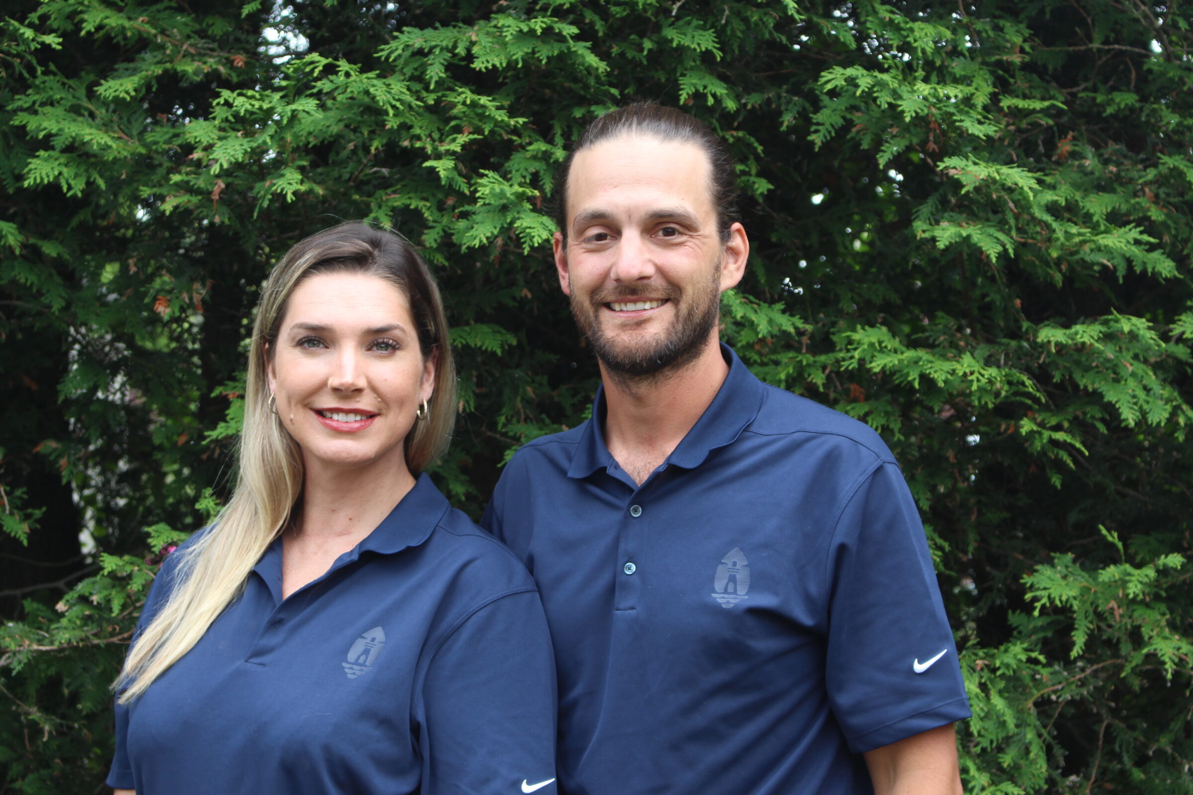 Two people stand in front of green foliage, smiling. They wear matching dark blue polo shirts with a logo, seeming professional and approachable.