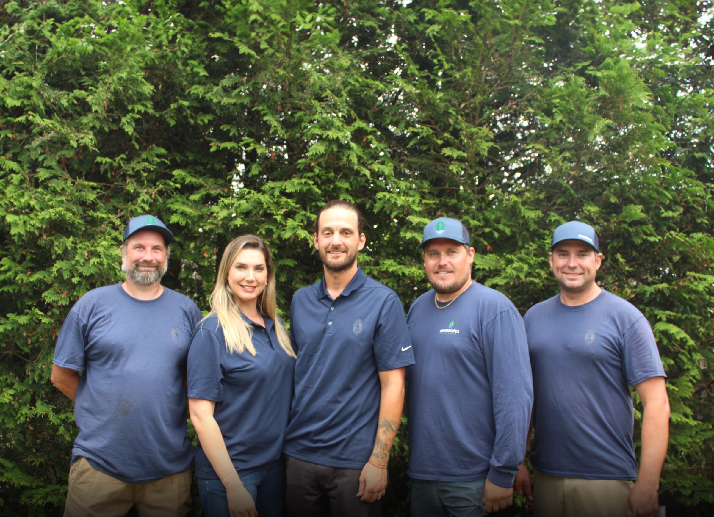 Five people wearing matching blue uniforms with caps stand smiling before a green leafy background, possibly a team or company staff photo.