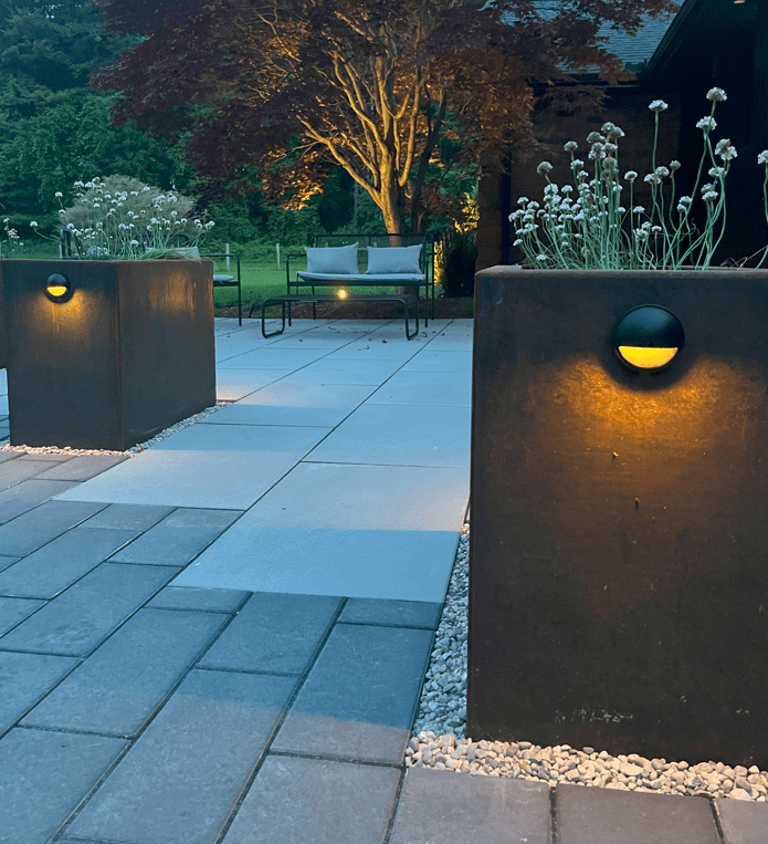 An outdoor space at dusk with large planters featuring illuminated cutouts, a bench, paved flooring, greenery in the background, and a tranquil ambiance.