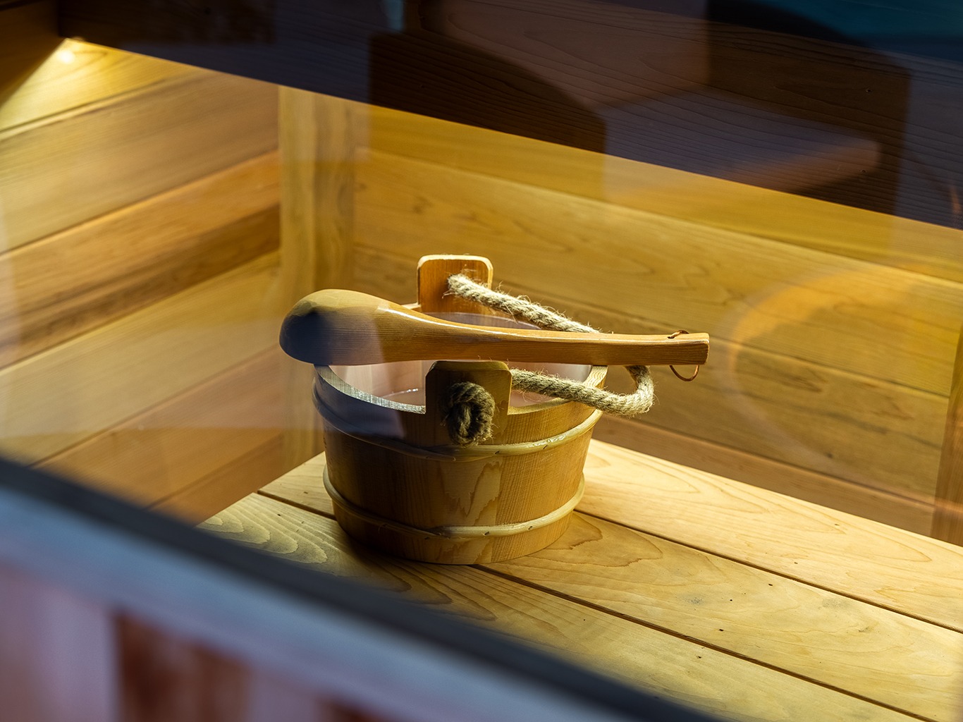 Close-up view through a glass window of a wooden sauna interior featuring a traditional wooden bucket with rope handle and matching ladle resting on a slatted bench. Warm interior lighting highlights the natural wood grain, with a faint reflection of a pool visible in the glass.