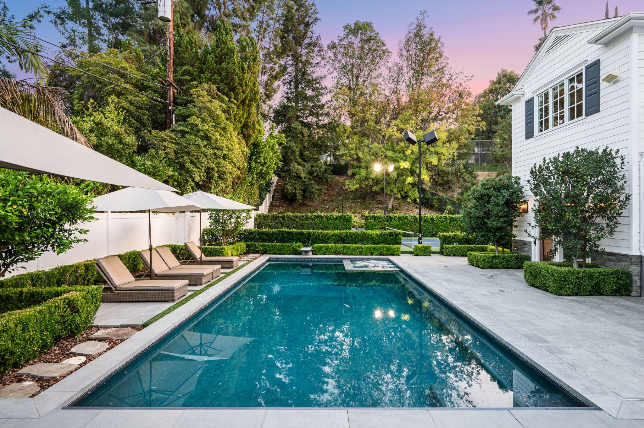 Rectangular swimming pool with stone patio, lounge chairs, and manicured hedges