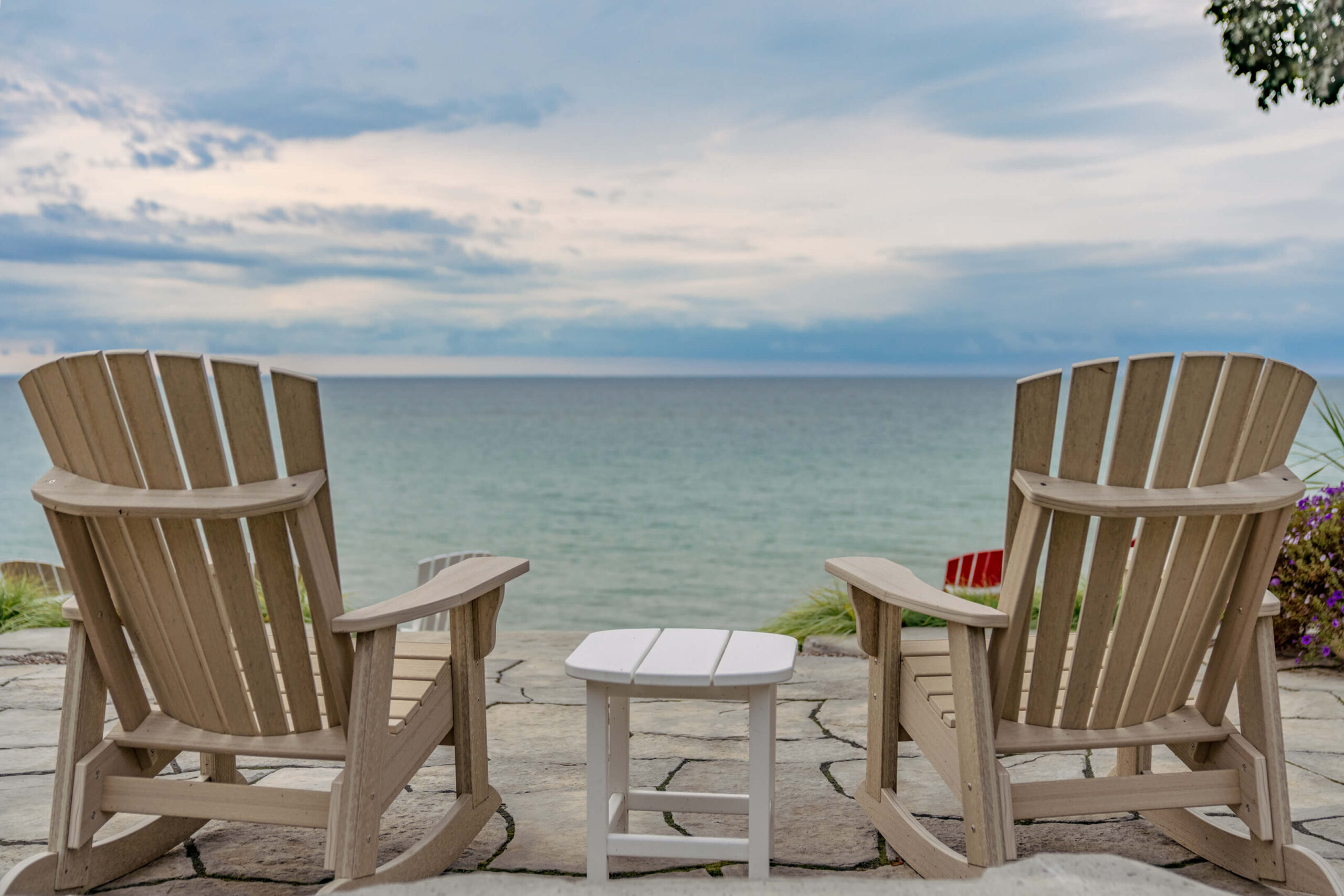 Two wooden Adirondack chairs sit on a stone patio overlooking a calm lakeshore, with a small white side table positioned between them. The chairs face the open water and a cloudy horizon, creating a peaceful waterfront seating area with an unobstructed view of the lake.