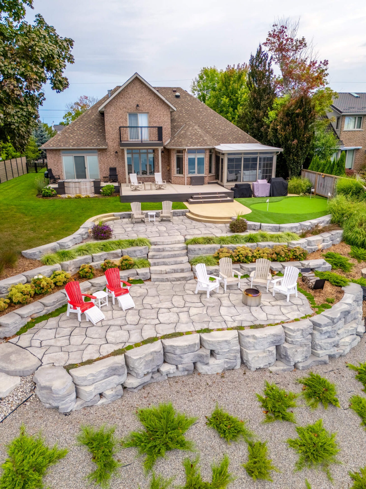 A large brick house with a tiered backyard patio featuring stone retaining walls, multiple seating areas with Adirondack chairs, a central fire pit, manicured landscaping, and a small putting green.