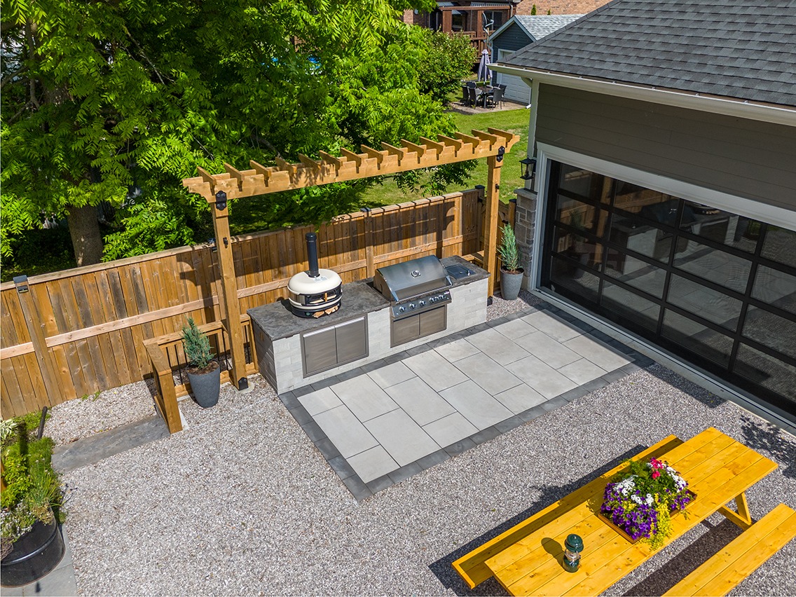 Aerial view of a backyard outdoor kitchen featuring a built-in stainless steel grill and countertop set into a stone base beneath a wooden pergola. The space includes a modern glass garage-style door on the home, gray patio pavers, decorative gravel ground cover, and potted plants. A bright yellow picnic table with a flower planter sits in