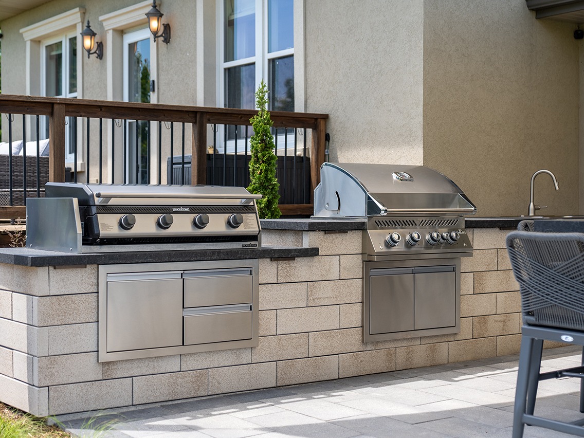Built-in outdoor kitchen featuring two stainless steel grills set into a light stone island with dark countertops and storage drawers below. A sink with a curved faucet sits on the right end of the counter. The setup is positioned against a home exterior with large windows, wall-mounted lantern lights, and a wooden railing, creating a functional backyard cooking and entertaining space.