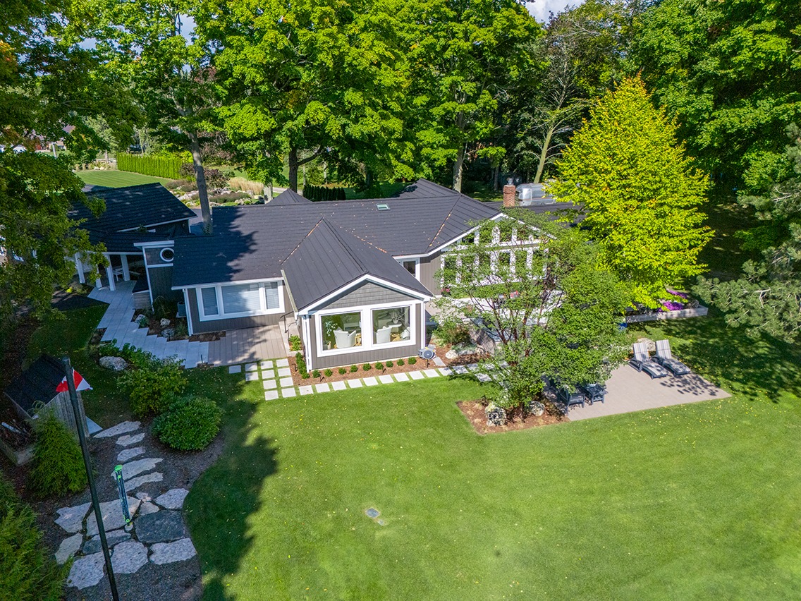 Aerial view of a landscaped backyard and single-story home with a dark metal roof surrounded by mature trees. A large, neatly manicured lawn stretches across the foreground, bordered by garden beds, stepping stone pathways, and a small patio seating area with lounge chairs. The home features large windows, light siding with decorative trim, and multiple connected outdoor living spaces nestled into lush greenery.