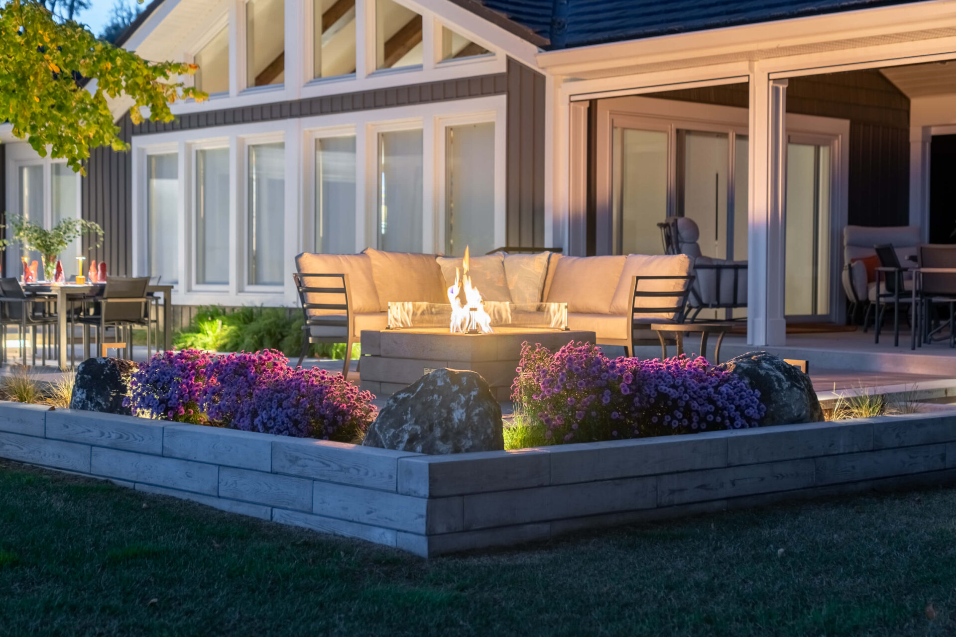 A modern backyard patio at dusk with a lit rectangular fire table, cushioned outdoor sofa seating, raised flower beds with purple blooms, and a contemporary home with large windows in the background.