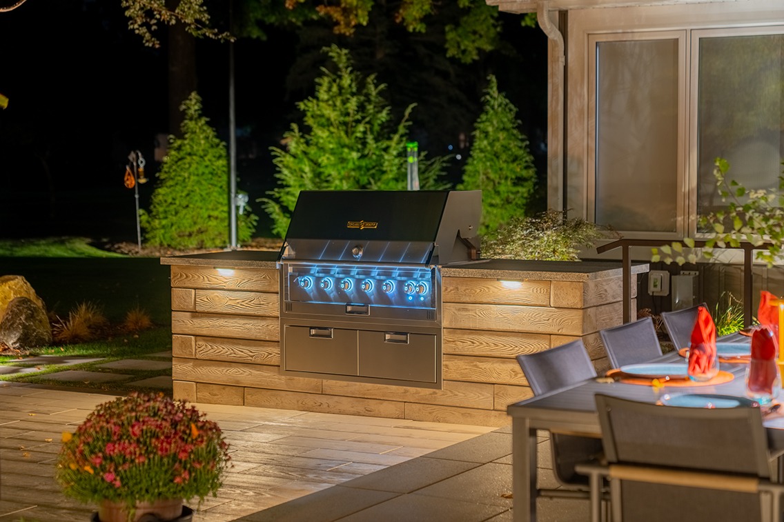 Nighttime view of a backyard outdoor kitchen featuring a built-in stainless steel grill with illuminated control knobs set into a wood-look stone island. Soft under-counter lighting highlights the textured façade, while landscaped greenery and pathway lights glow in the background. In the foreground, a dining table with chairs and red napkins creates an inv