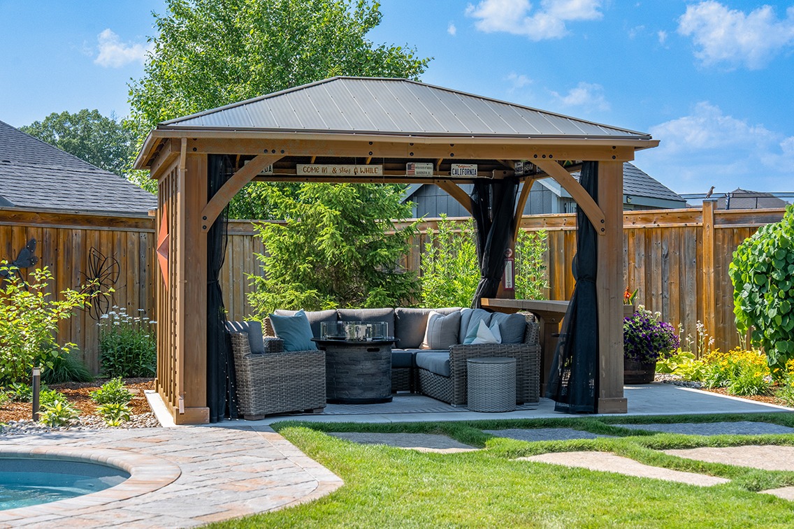 Bright daytime view of a backyard gazebo with a metal roof and wooden posts, featuring black privacy curtains and a wicker sectional sofa arranged around a round fire table. The gazebo sits on a concrete patio surrounded by landscaped garden beds, a wooden privacy fence, and lush greenery. In the foreground, a manicured lawn with stepping stones leads toward the edge of a curved in-ground pool.