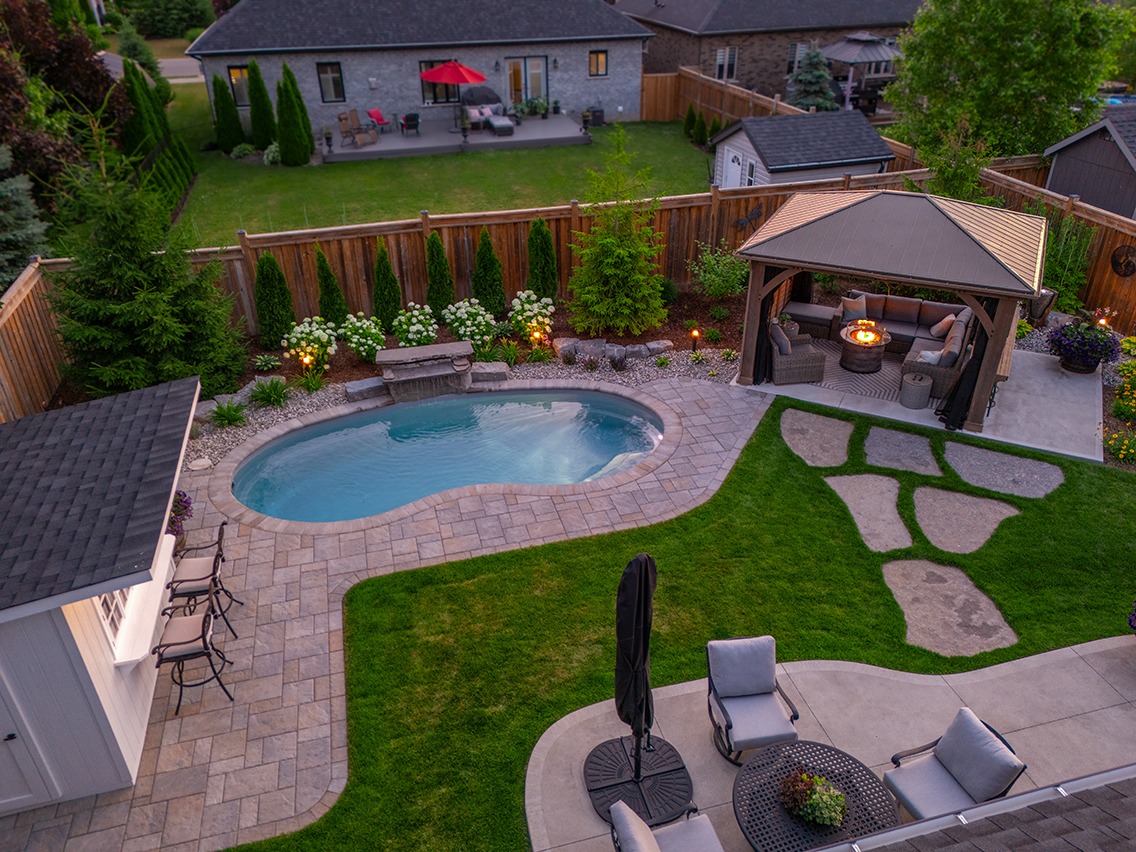 Aerial view of a landscaped backyard at dusk featuring a curved in-ground pool surrounded by stone pavers and garden beds with shrubs and accent lighting. A covered gazebo with seating and a glowing fire table sits beside a manicured lawn with large stepping stones. Wood fencing, neighboring homes, and a small shed frame the inviting outdoor living space.