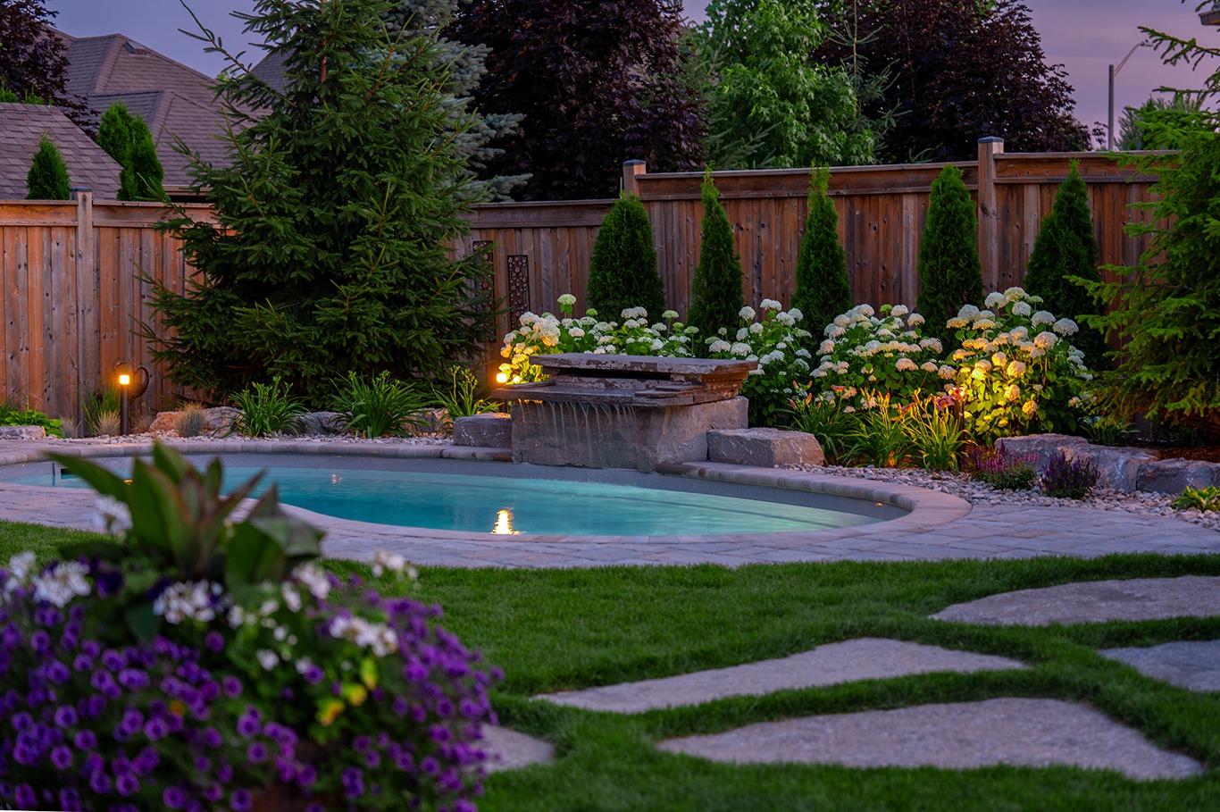 Evening view of a curved backyard pool with a stone waterfall feature cascading into the water. Landscape lighting illuminates flowering plants, shrubs, and tall evergreens along a wooden privacy fence. In the foreground, stepping stones cross a lush green lawn with purple flowers blooming nearby.