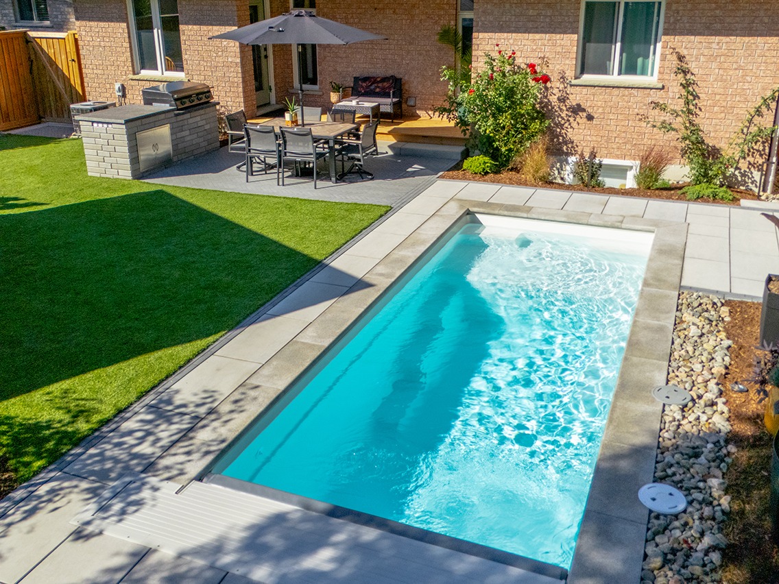 Backyard view of a bright blue rectangular pool, stone patio, and a brick house with an outdoor dining area.