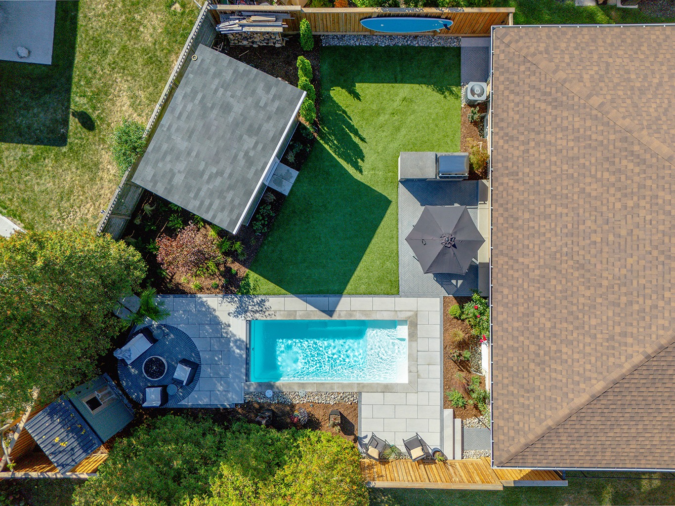 Top-down aerial view of a modern backyard with a rectangular in-ground pool bordered by large gray stone pavers. A covered patio structure sits on one side, with a neatly trimmed lawn, wood fencing, and a small seating area with an umbrella adjacent to the house. Clean landscaping lines and geometric design elements define the space.