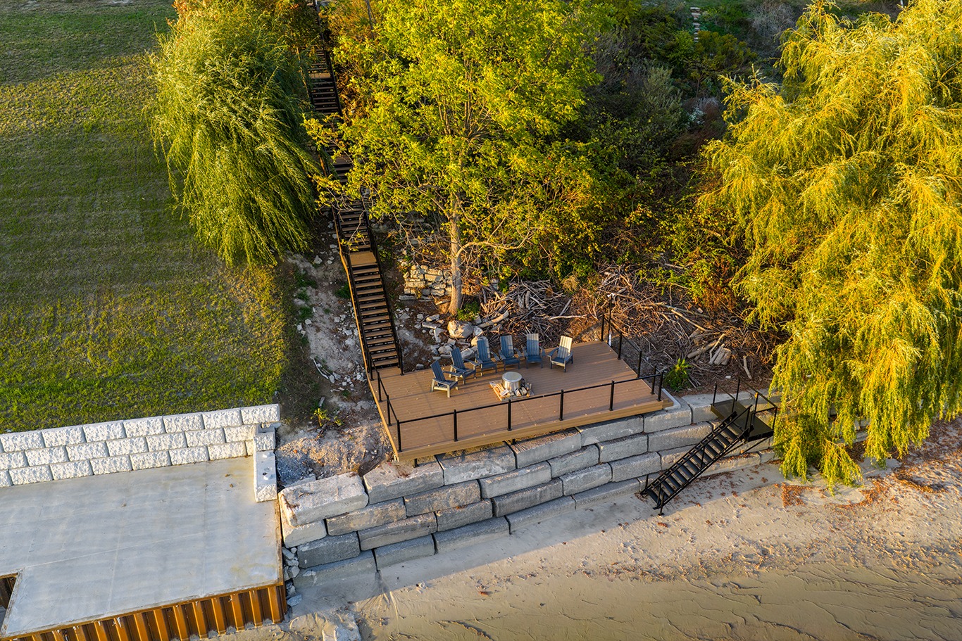 Aerial view of a waterfront backyard deck built on a stone retaining wall, featuring four blue Adirondack chairs around a small fire pit table. Black metal stairs lead up the landscaped slope to the lawn above, surrounded by mature trees with golden evening sunlight filtering through the leaves. A sandy shoreline sits below the deck.