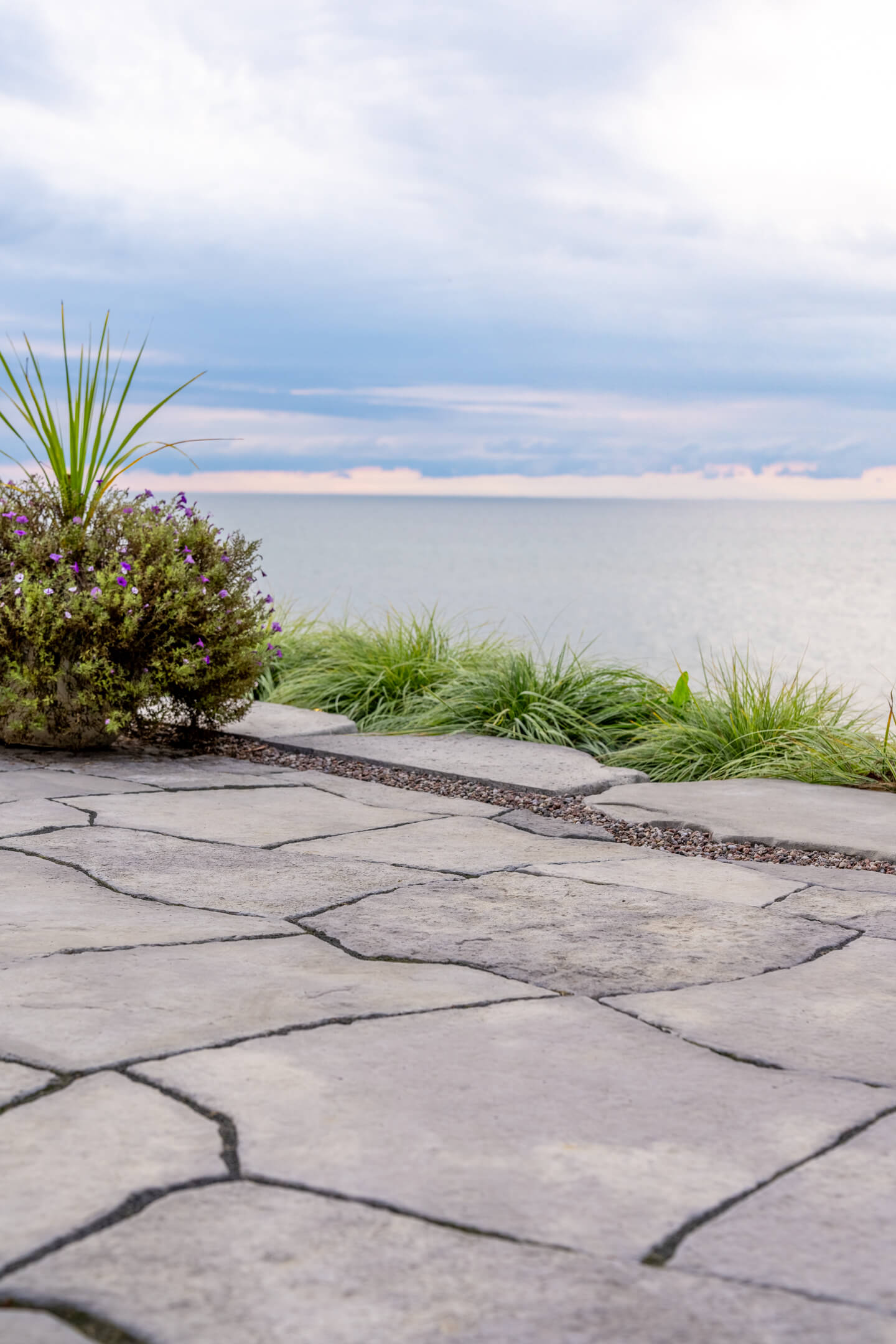 Low-angle shot of a flagstone patio bordered by green grass and a purple flower planter near the water.
