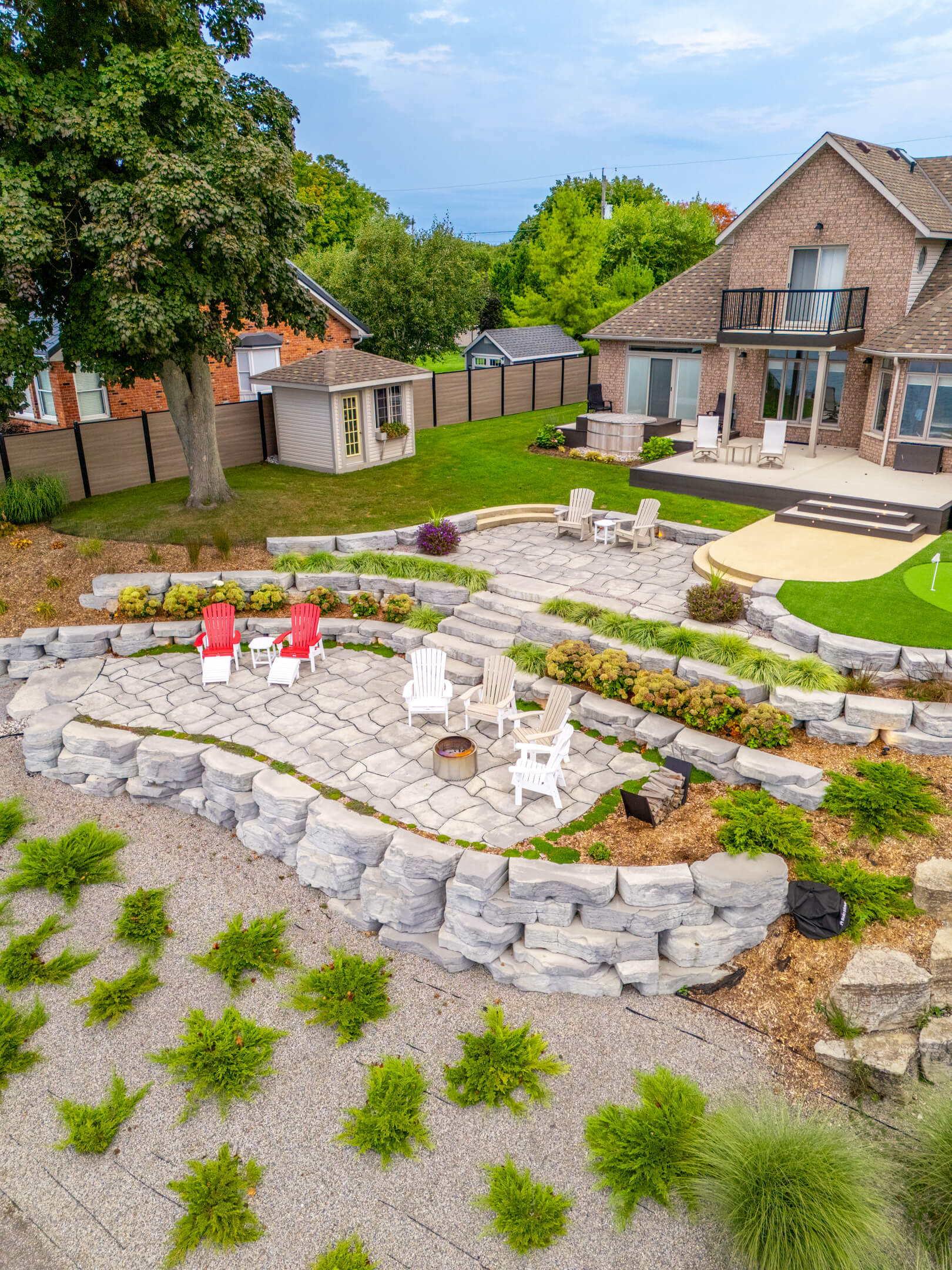 Wide aerial shot of a landscaped backyard featuring stone terraces, a shed, and seating around a fire pit.