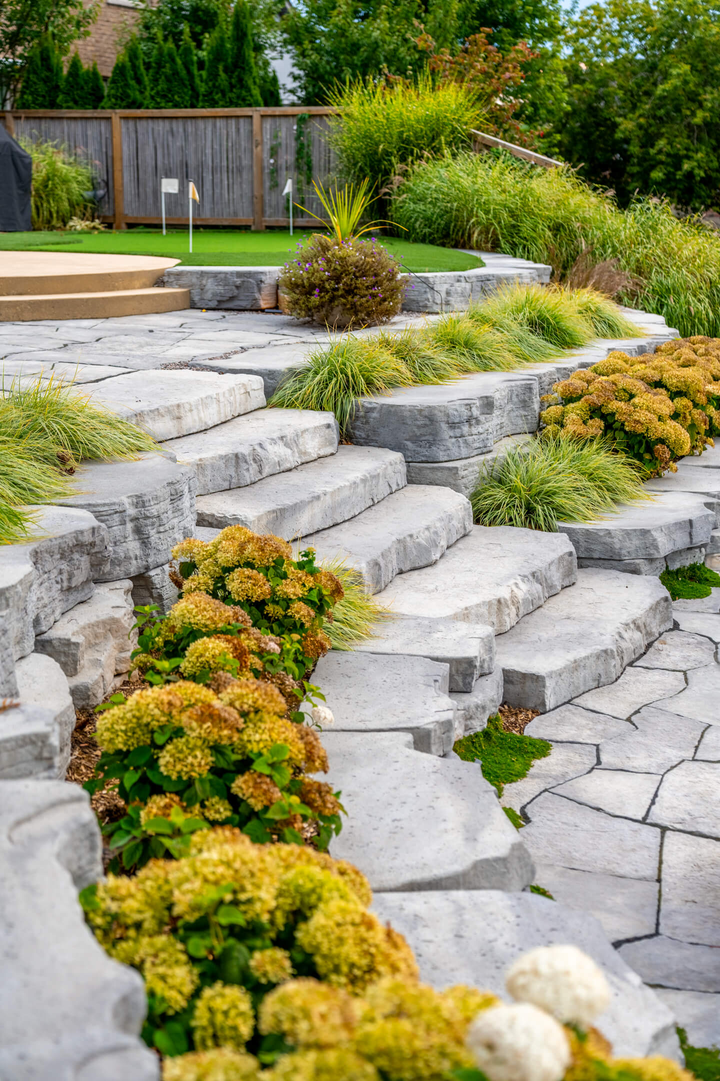 Tiered backyard landscaping with large stone steps, hydrangeas, and a small putting green in the background.