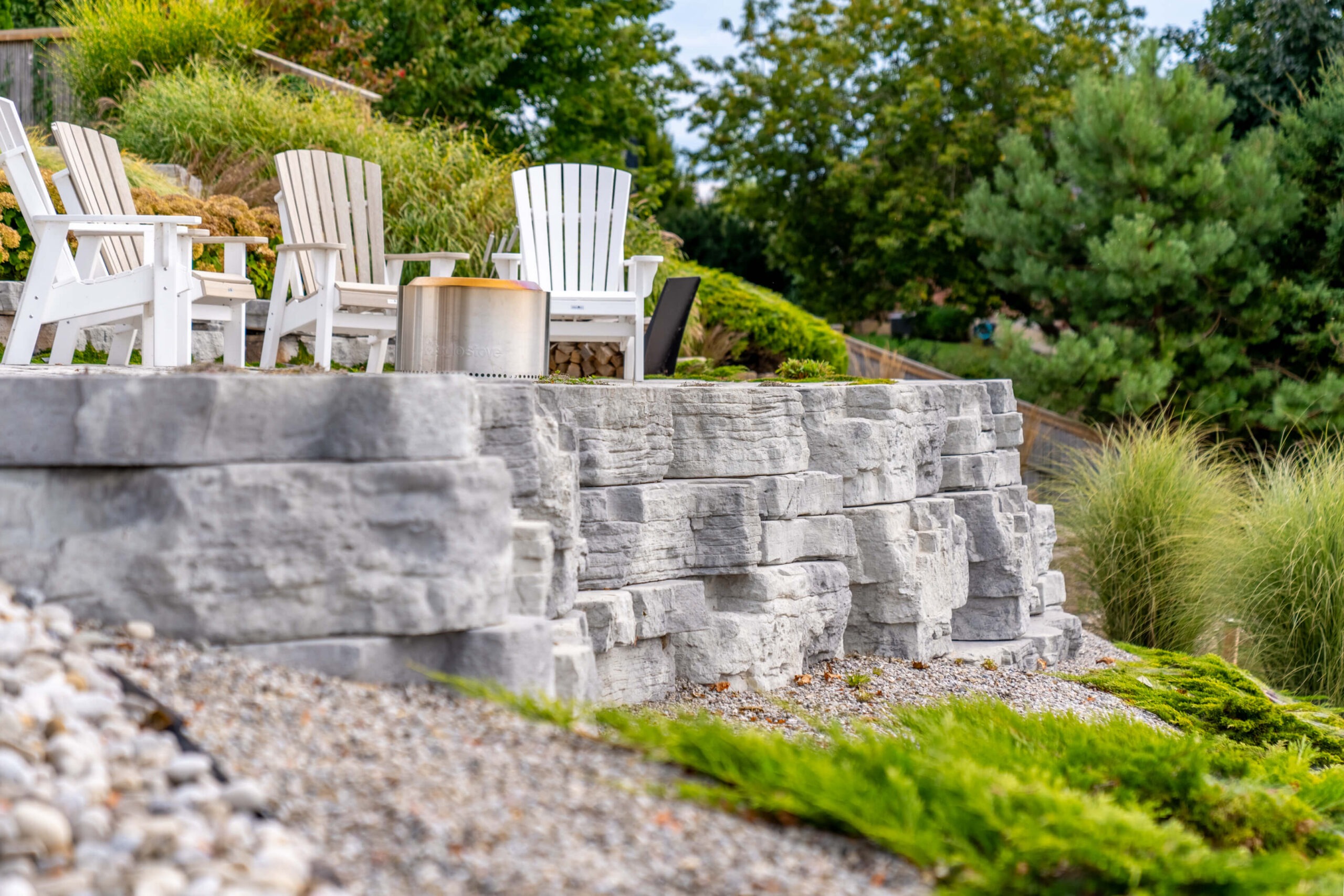 Profile of a stacked stone retaining wall with Adirondack chairs and a metal fire pit on the upper terrace.