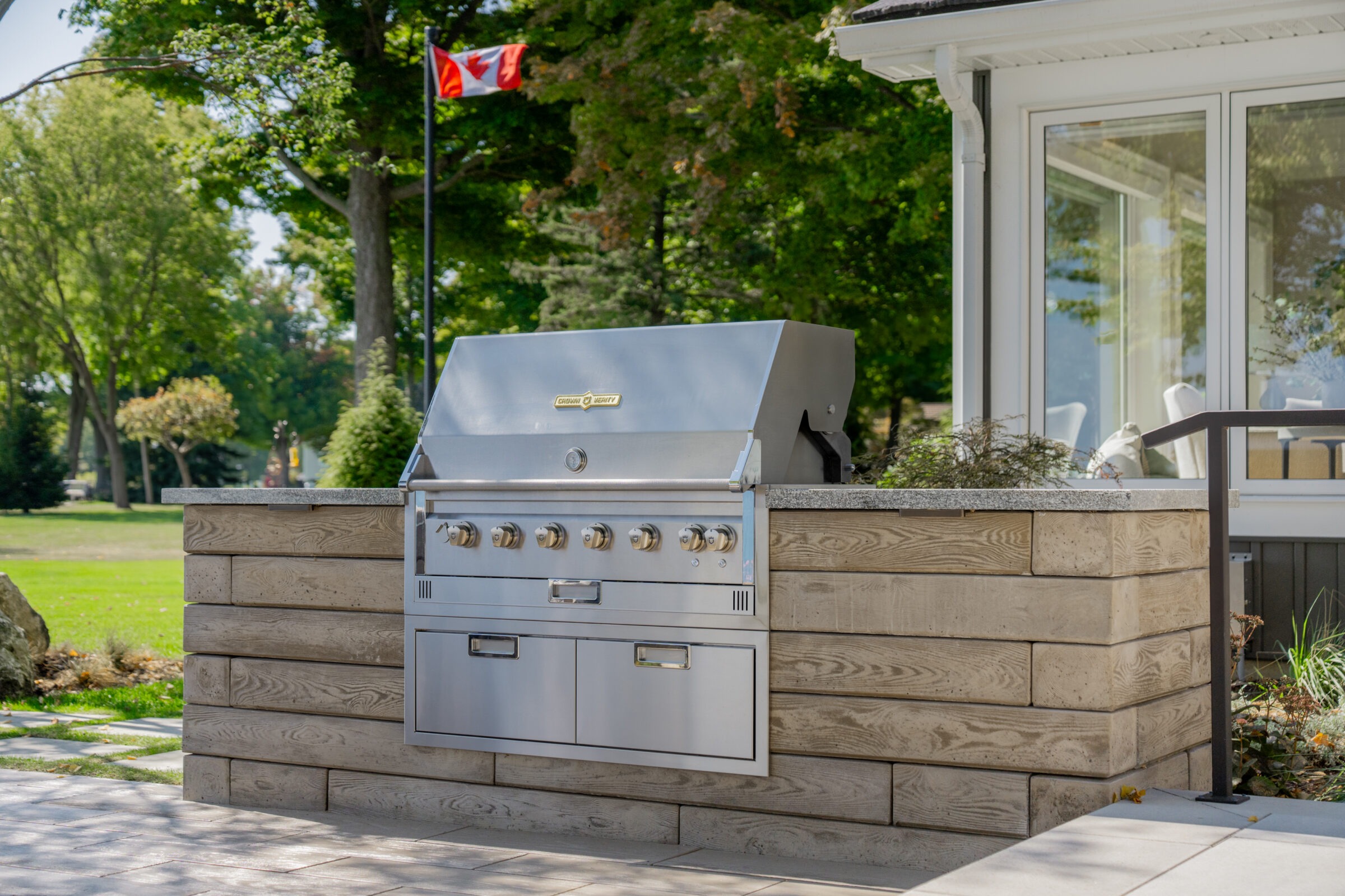 : Front view of a built-in stainless steel grill station on a stone patio with a Canadian flag in the background.