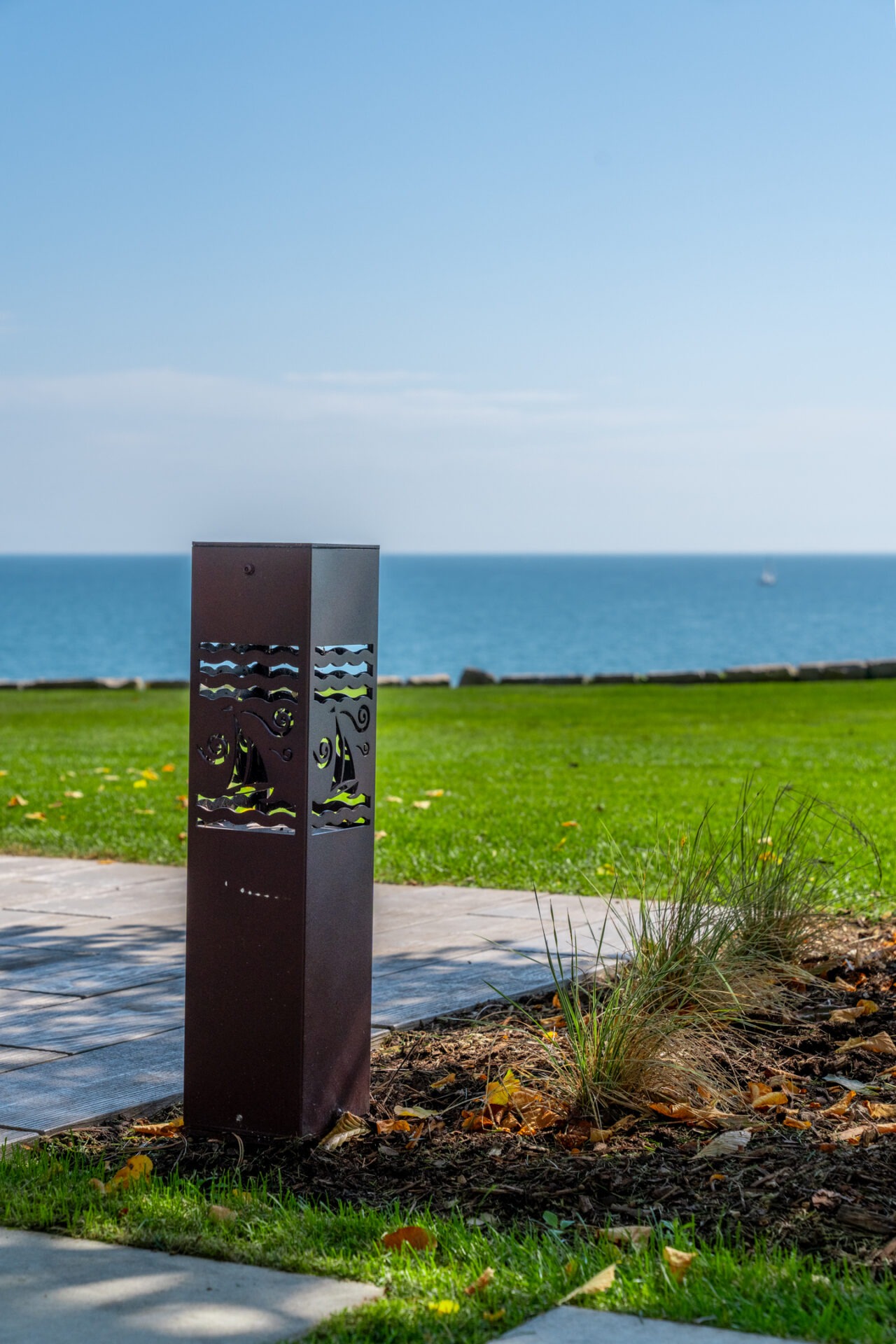 A decorative metal bollard light with sailboat cutouts stands in a garden bed overlooking the lake.