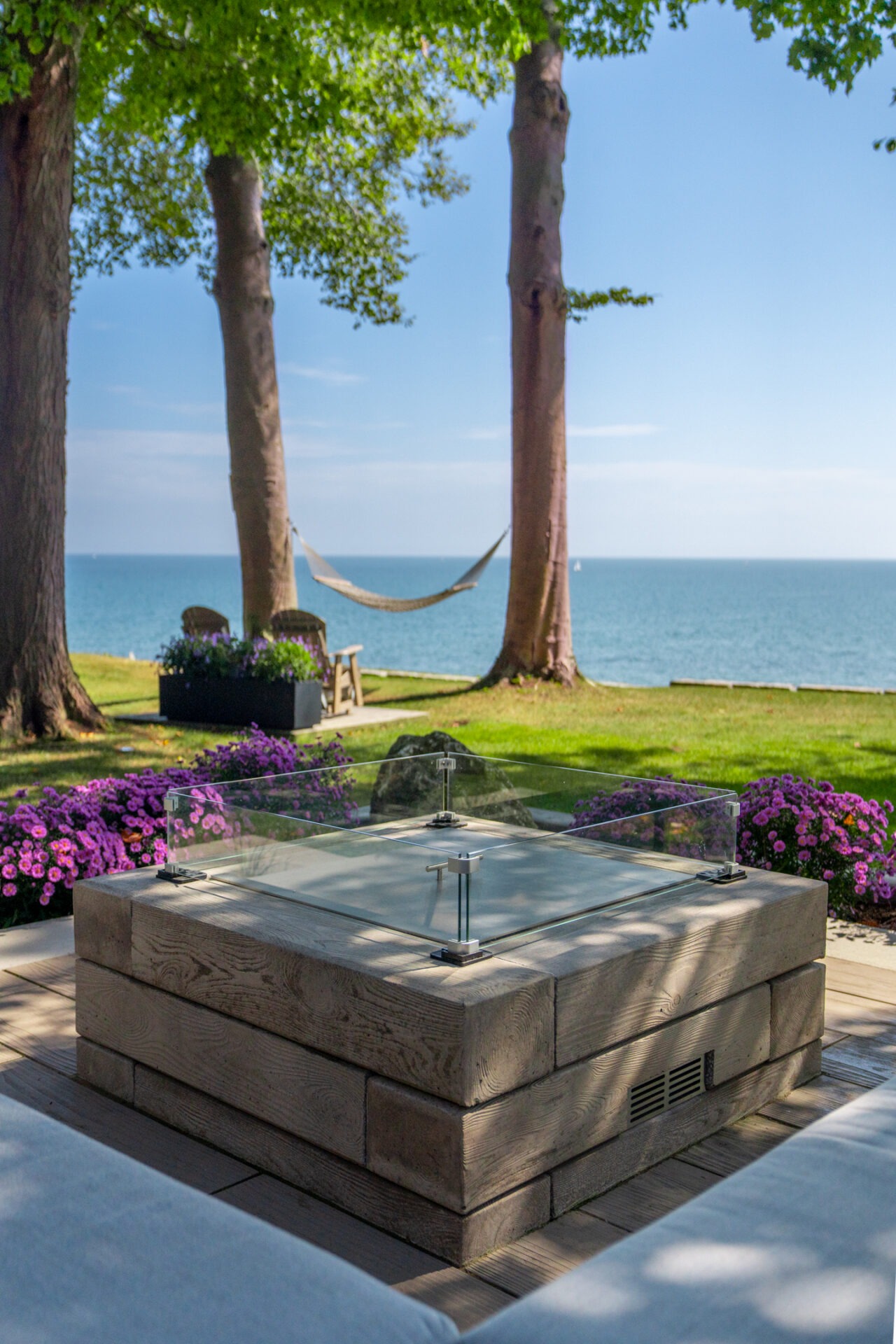 A square fire pit with a glass wind guard on a stone patio, with a hammock and lake in the background.
