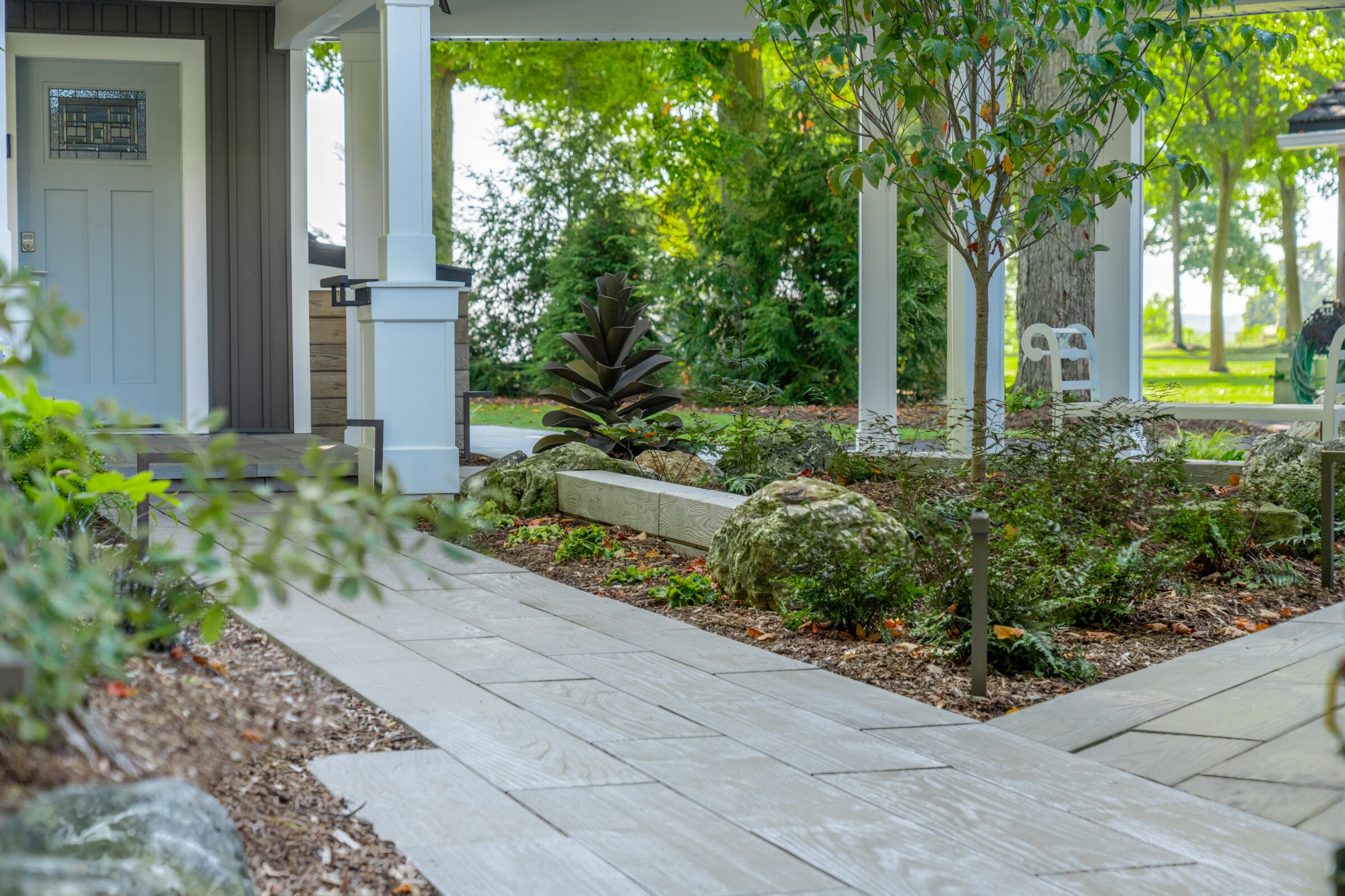 A wood-plank stone path leading through a landscaped garden area with white pillars and a metal sculpture.