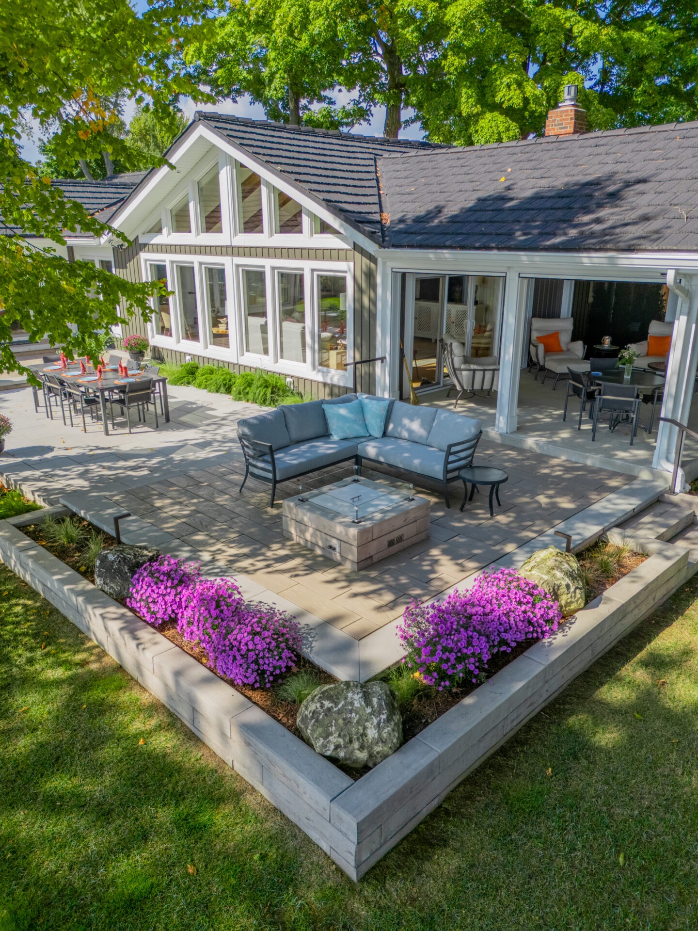 Bird's-eye view of a tiered patio with a sectional sofa, fire pit, and purple flowers near a modern home.