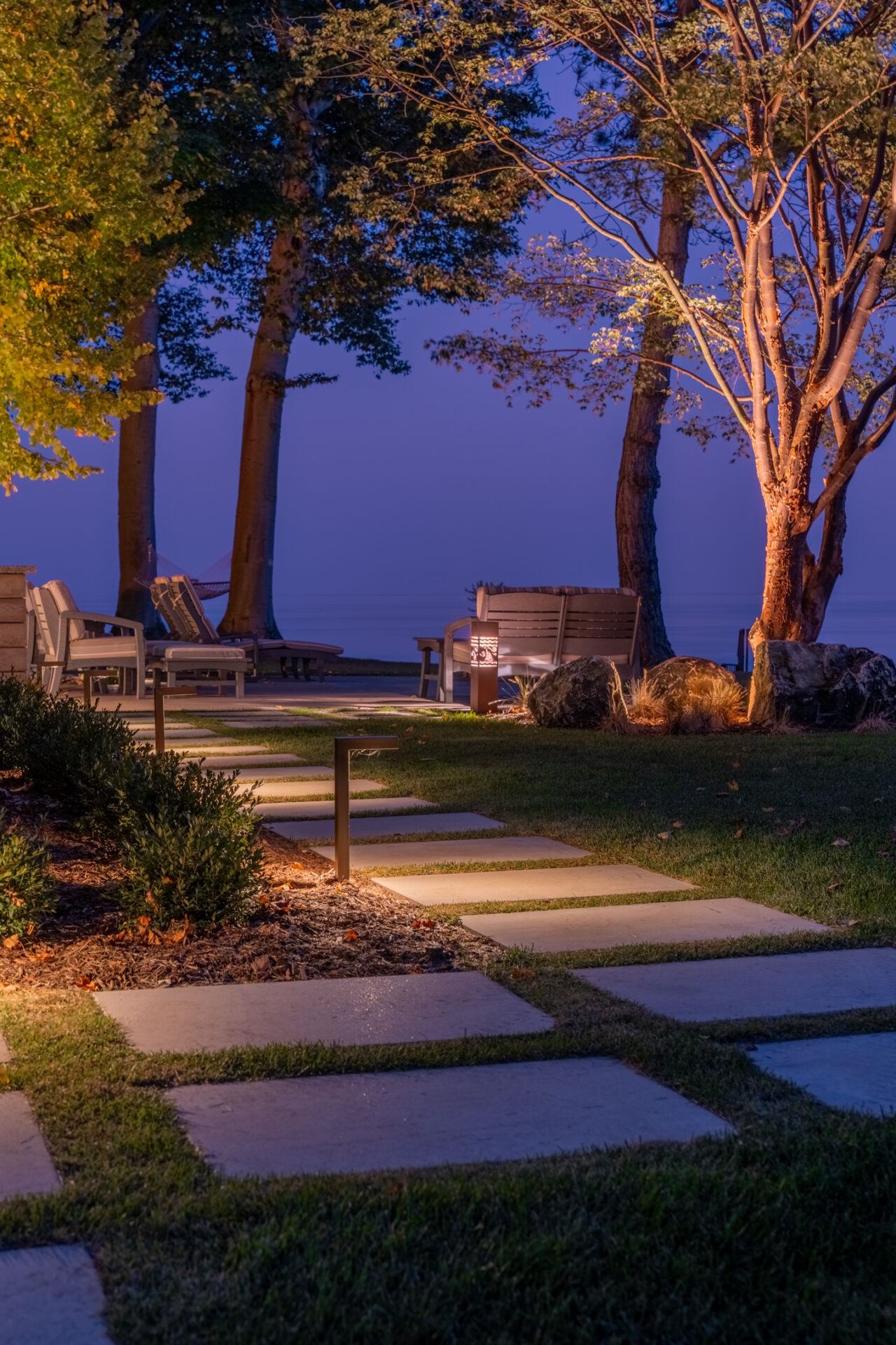 Evening view of a stone paver path lit by bollard lights leading toward a calm lake at twilight.