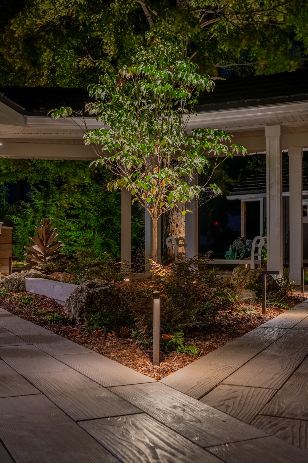 Close-up of a decorative garden area at night with a lit tree and stone pathways under soft warm light.