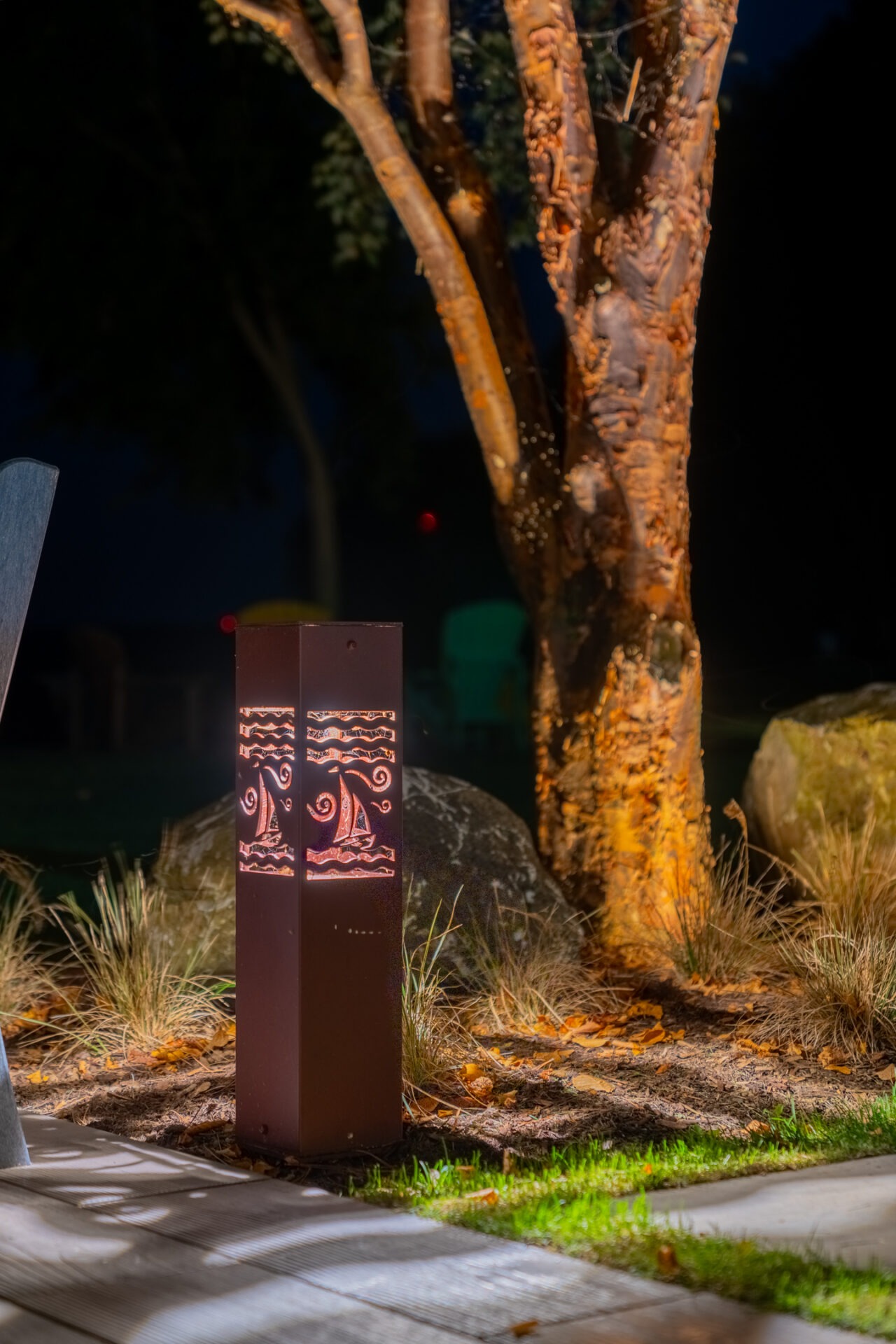 A lit decorative bollard with sailboat cutouts glowing in a garden bed near a tree at night.
