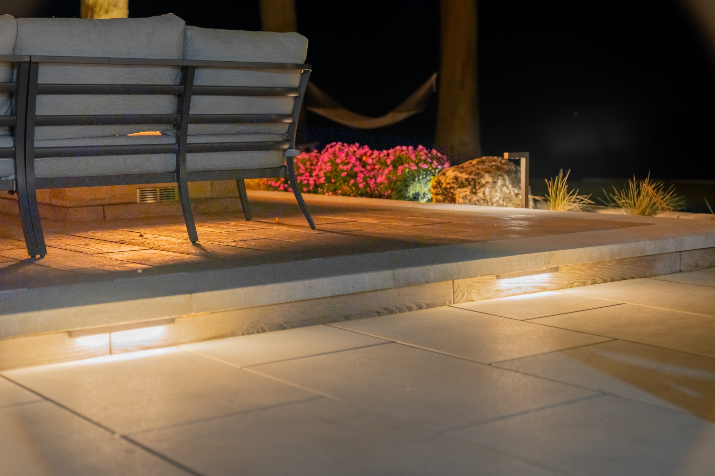 Night view of a stone patio with integrated step lighting, a sectional sofa, and glowing garden beds.